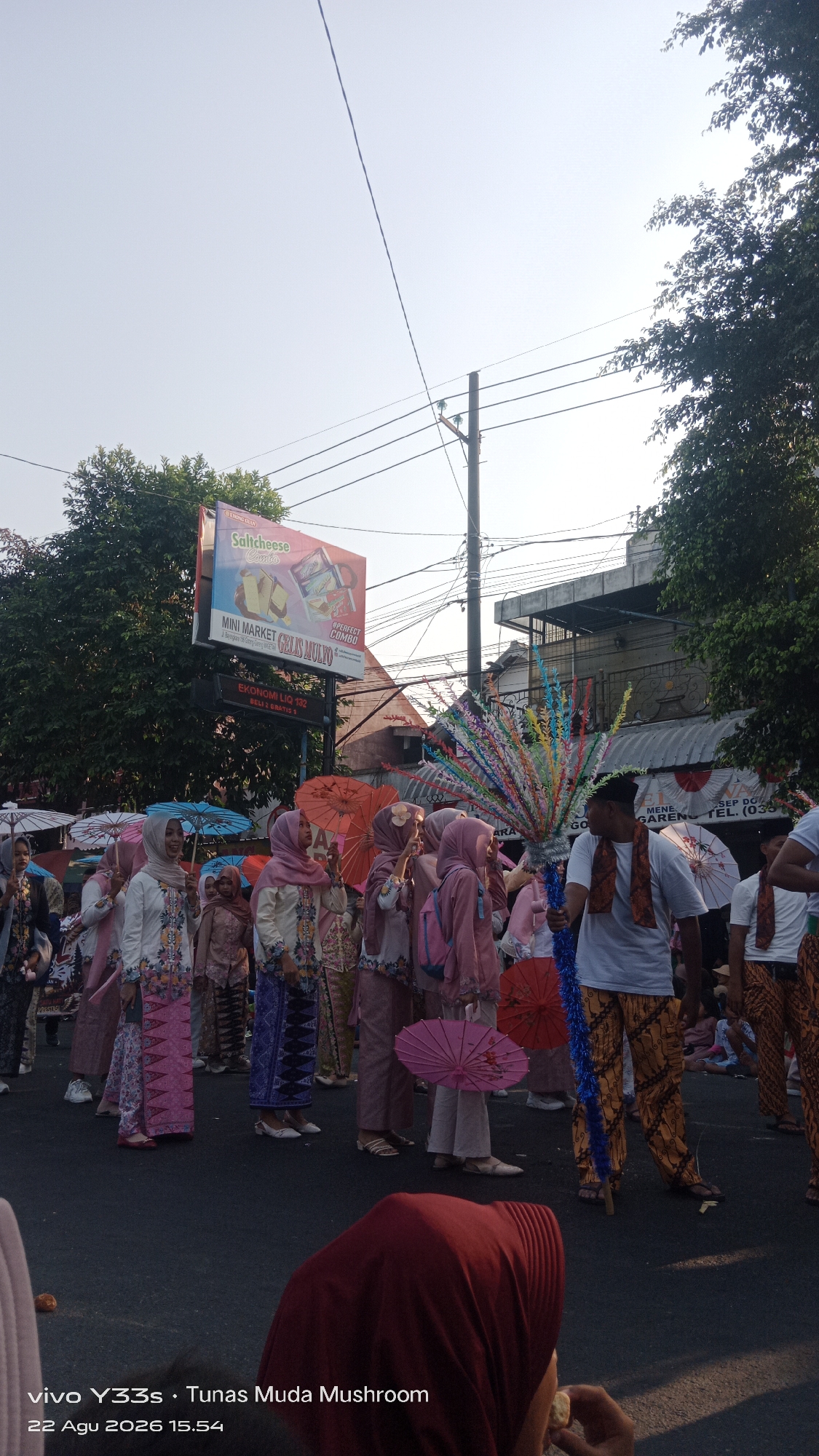 A group of people, primarily women, are participating in a parade on a street in Genengan, Indonesia. They are dressed in traditional Indonesian attire, including headscarves and patterned sarongs. Many are holding colorful, decorative umbrellas. A man in the foreground holds a staff adorned with colorful streamers. In the background, a billboard advertises "Salicheese Combo" and "Mini Market Gelis Mulyo." The scene appears to be taking place during the daytime, with a clear sky and trees lining the street. The ground is paved, and there are buildings and utility poles visible. The timestamp "22 Agu 2026 15:54" is superimposed on the lower left corner of the image.