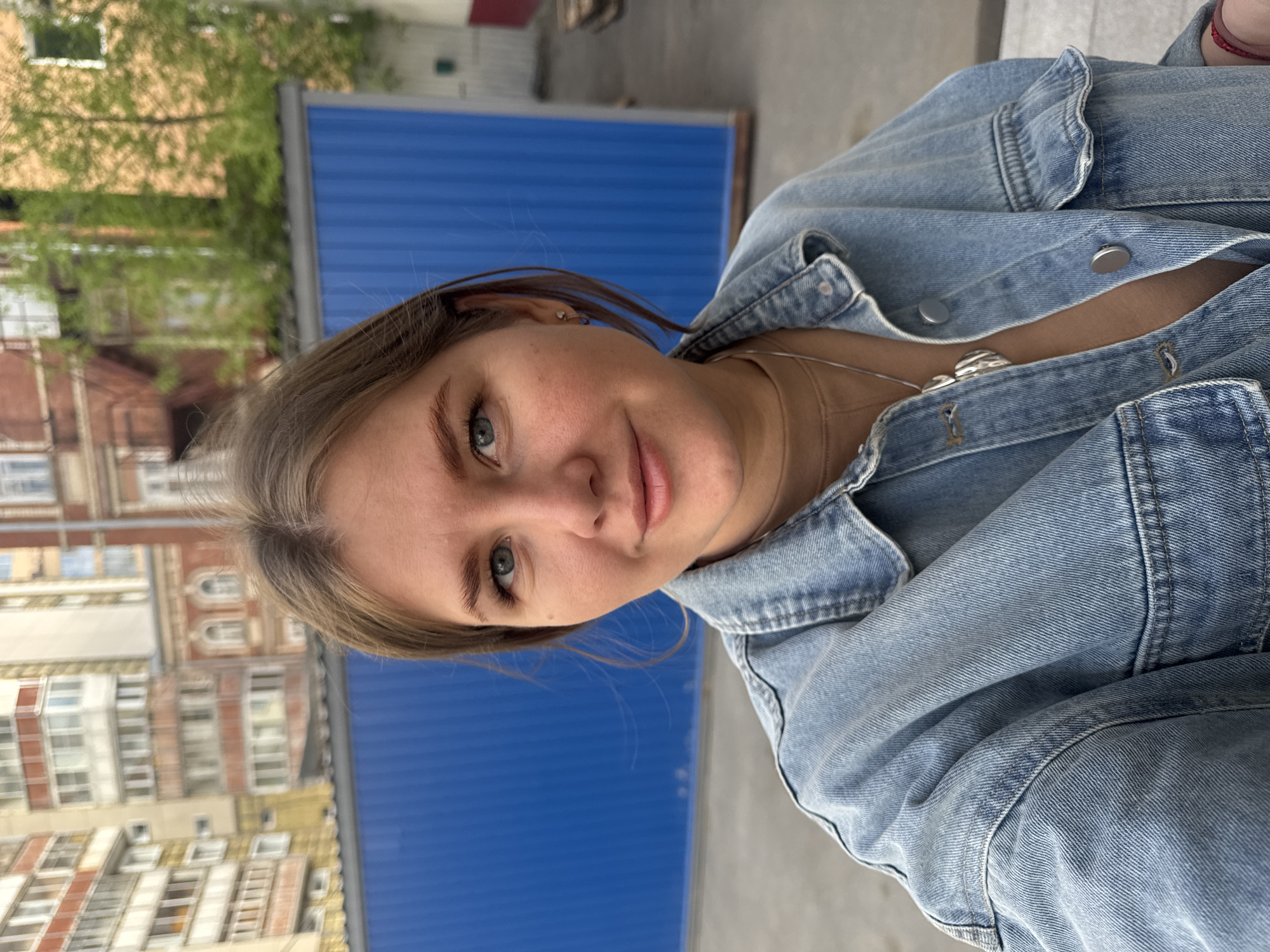 A young woman with fair skin, light brown hair, and green eyes is wearing a light blue denim jacket and a silver necklace. She appears to be outdoors in Perm, Russia. Behind her, there is a blue corrugated metal fence and buildings with distinct architectural features characteristic of Russian urban areas. Some foliage is also visible. The woman is facing the camera with a slight smile.