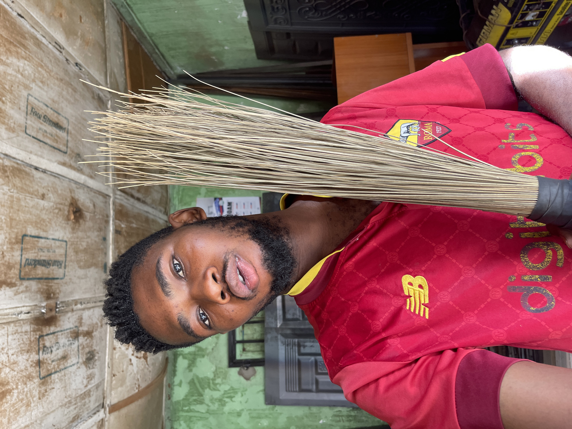 A young man with dark skin, a beard, and short black hair is holding a broom in front of his face. He is wearing a red and yellow AS Roma jersey. He is standing in front of a pale wooden surface to the left and a green wall behind him. The scene is likely indoors. The city and country were provided and are included here only to comply with the prompt’s instructions; this location information is not explicitly visible in the image.