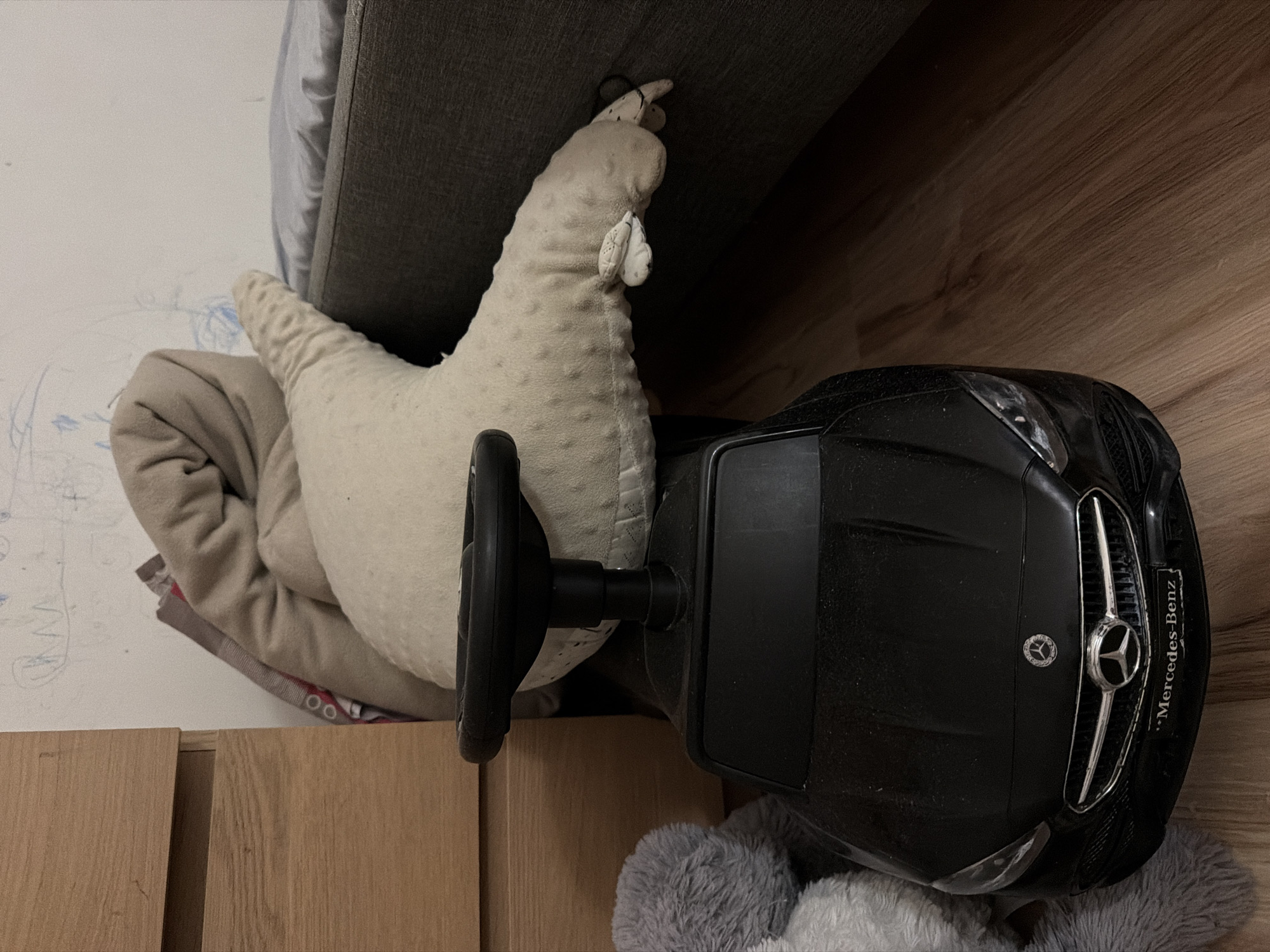 A black toy Mercedes-Benz car is positioned on a wooden floor. A plush, light-colored crescent-shaped pillow rests on top of the car, partially obscuring its steering wheel. To the left of the car, a portion of a gray stuffed animal is visible. In the background, a wooden dresser is on the left, and a bed with a dark gray headboard and light blue bedding is on the right. The wall behind the objects appears to be white with some faint, crayon-like drawings. The lighting in the scene is dim. This image was taken in Falenty Nowe, Poland.