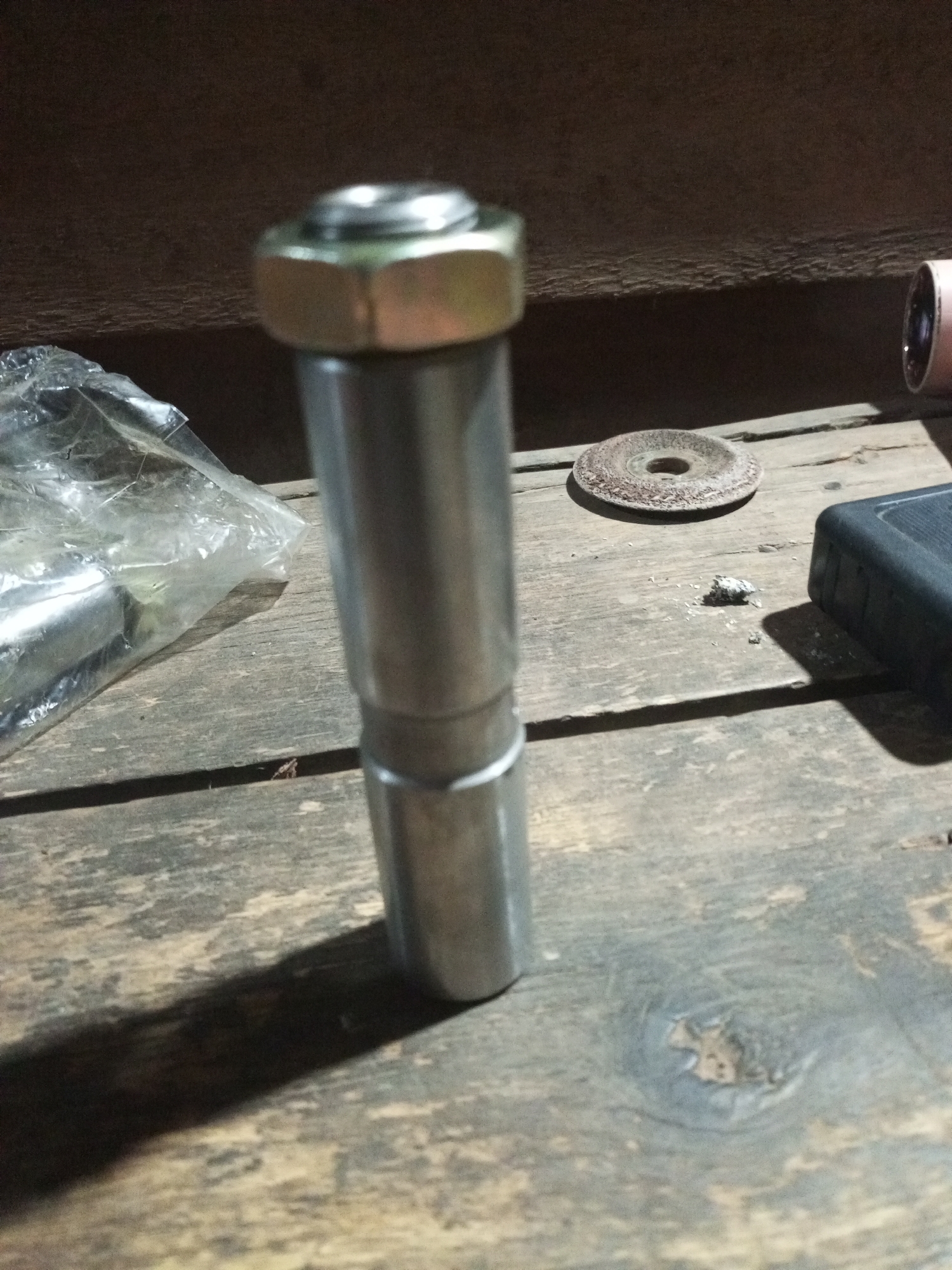 The image shows a close-up, overhead view of a metallic object with a nut on top, resting on a wooden surface. The object is cylindrical and appears to be a bolt or a similar fastener. To the left of the object, a clear plastic bag containing more metallic items is visible. In the background, a dark surface can be seen, likely a wall or shelf. The wooden surface appears weathered and has visible grain and knots. There are also a few small debris pieces and a circular abrasive disc on the surface. The lighting suggests an indoor setting, possibly a workshop or storage area, with harsh shadows cast by the objects. There are no people or animals present in the image. The location is described as Unknown, Unknown.