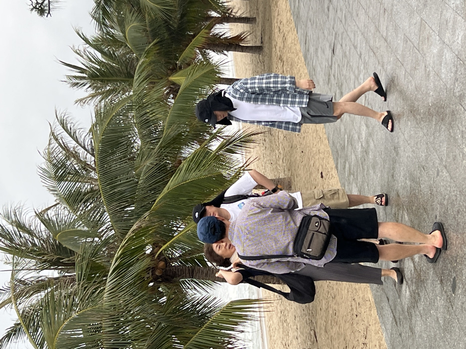 The image depicts a beachfront scene in Nha Trang, Vietnam, featuring a paved walkway adjacent to a sandy beach lined with palm trees. The sky is overcast, and the paved walkway appears damp.
Four individuals are present on the paved walkway.
1. In the foreground, an adult male stands with his back mostly to the camera, wearing a blue baseball cap, a light-colored striped collared shirt, black shorts, and black flip-flops. He carries a two-tone olive green and black messenger bag across his chest. He faces the other two individuals.
2. To his left, an adult female wears a dark top, dark wide-leg pants, and dark flat shoes, carrying a black shoulder bag. Her right hand is raised near her ear, and she appears to be speaking towards the center group.
3. Behind the foreground male, another adult male wears a black baseball cap, a white t-shirt with a light graphic, khaki cargo shorts, and dark flip-flops. He carries a white backpack and is part of the conversational cluster.
4. To the right of the foreground male, a fourth adult male is walking from left to right along the walkway. He wears a black bucket hat with a neck flap, a white t-shirt, an open blue and white plaid long-sleeved shirt, gray athletic shorts with a Nike logo, and black slide sandals.
The sandy beach, visible to the left, extends to calm ocean water. Several mature palm trees with visible coconuts line the demarcation between the sand and the paved area.