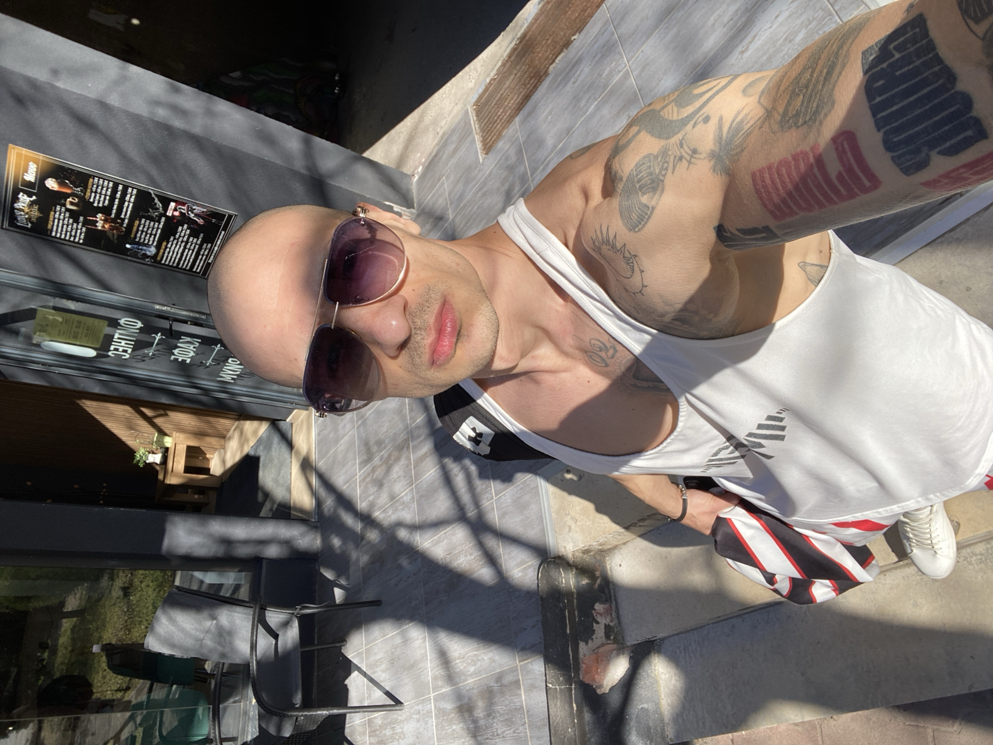 A bald man with sunglasses and tattoos on his arm is taking a selfie outside a cafe in Pleven, Bulgaria. He is wearing a white tank top and white sneakers, and he is holding a red and black striped towel. The cafe has a sign in Bulgarian.