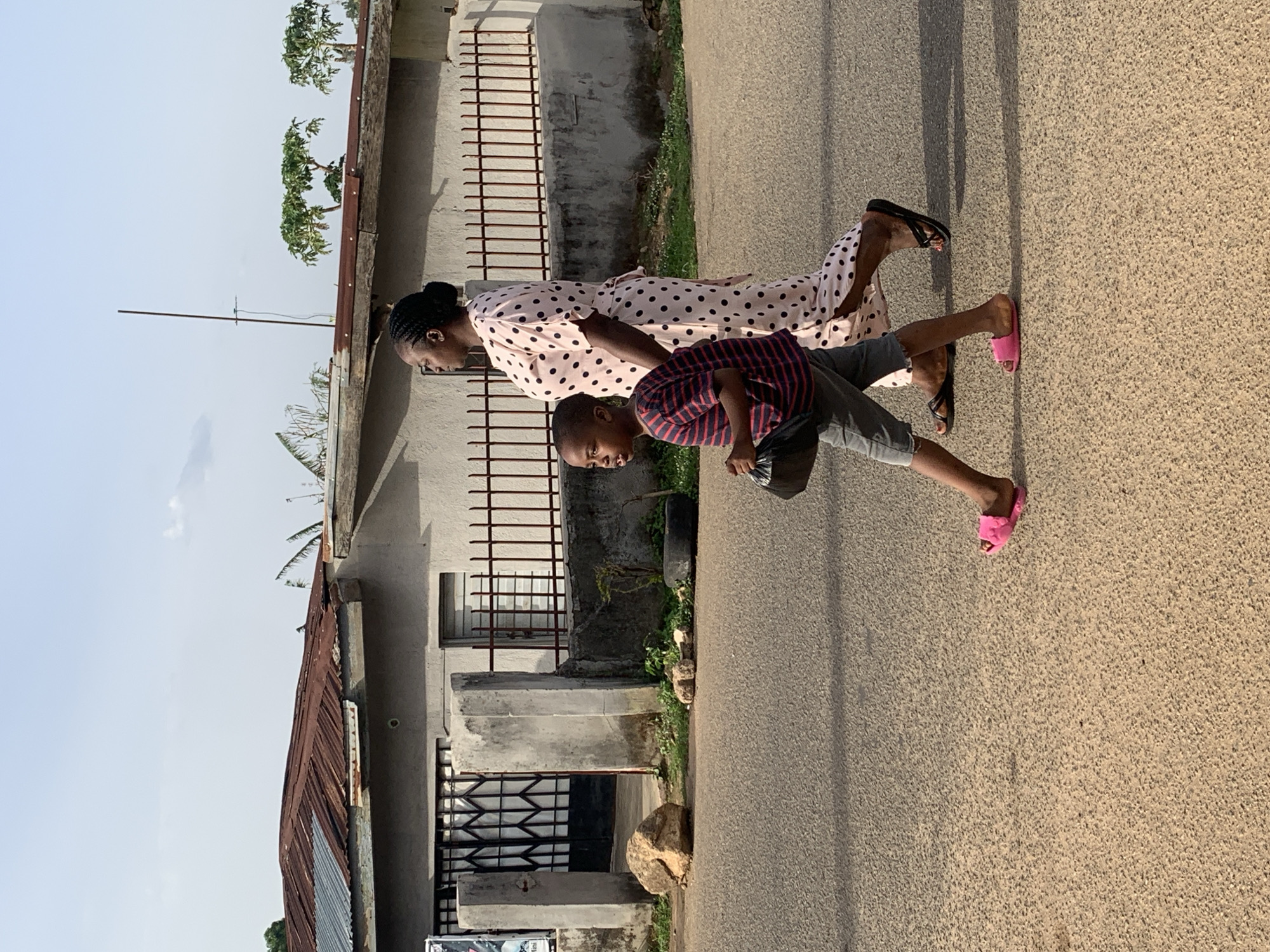 A woman and a boy are walking on an asphalt road in Jos, Nigeria. The woman is wearing a light pink polka-dot dress, and black sandals. The boy is wearing a striped red and black shirt, gray shorts, and pink sandals. They are walking past buildings with corrugated metal roofs and barred windows. The sky is overcast.