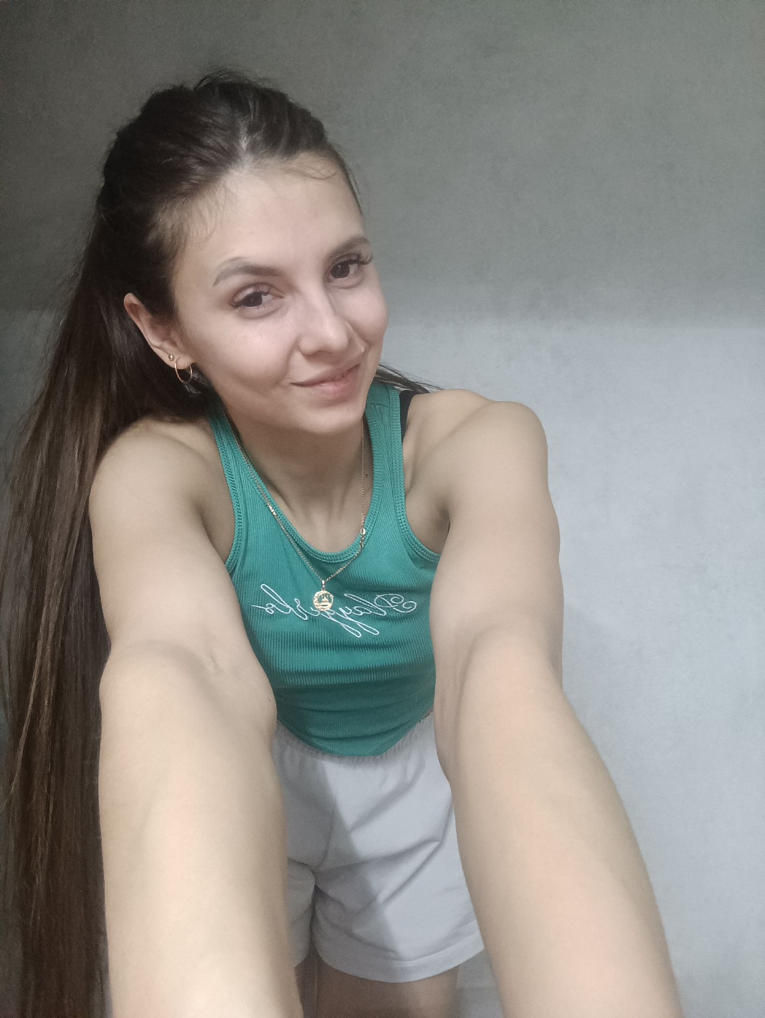 A young woman is taking a selfie indoors. She has long brown hair pulled back at the front, and is wearing a green tank top, a gold necklace, and white shorts. The background is a plain, light-colored wall. The location is Unknown, Unknown.