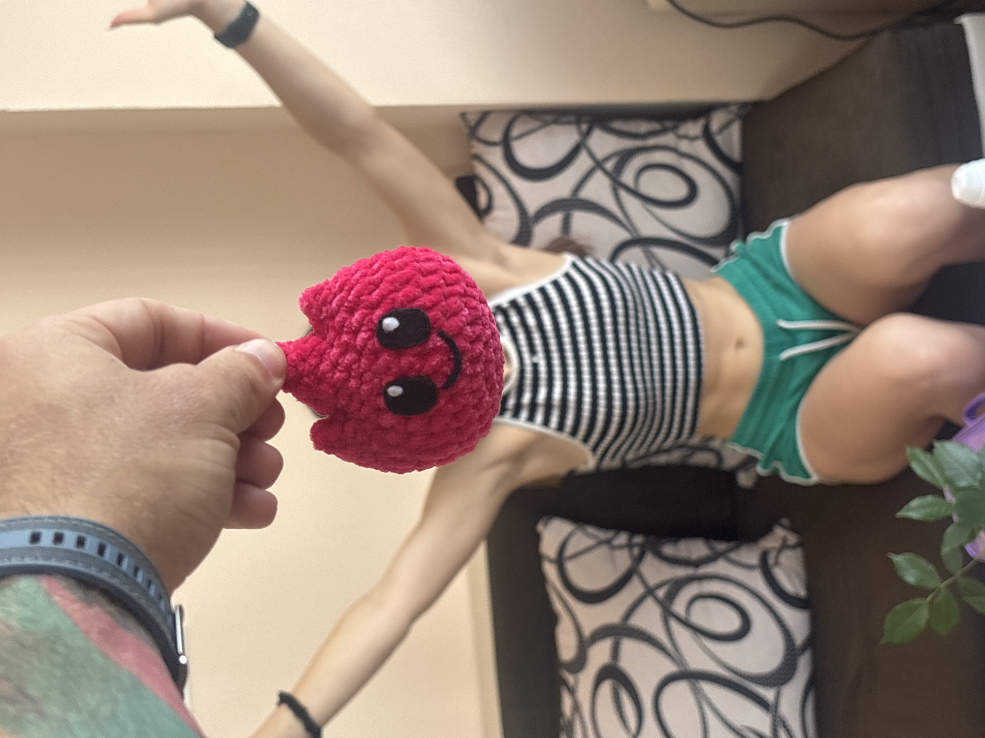 A hand with a smartwatch and tattoos holds a crocheted pink doll with a smiling face in front of a young woman sitting on a couch. The woman is wearing a black and white striped crop top and green shorts. She has her arms raised in the air. The background shows a wall and pillows on the couch. The scene is indoors, likely in a residential setting in Varna, Bulgaria.