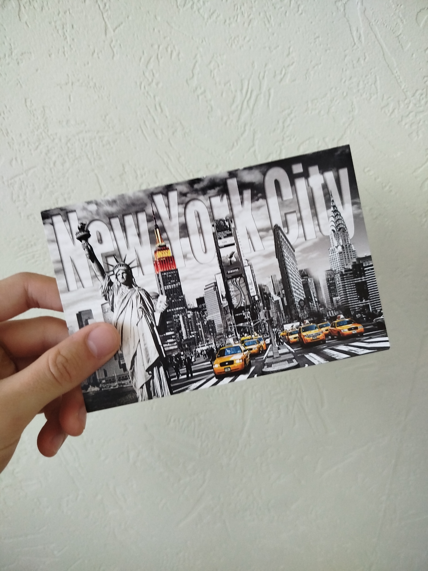 A hand holds a postcard featuring a black-and-white cityscape of New York City. Prominently displayed are the Statue of Liberty, the Empire State Building, Times Square, the Flatiron Building, and the Chrysler Building. Yellow taxis are visible on the street. The words "New York City" are superimposed over the image. The background is a textured off-white surface.