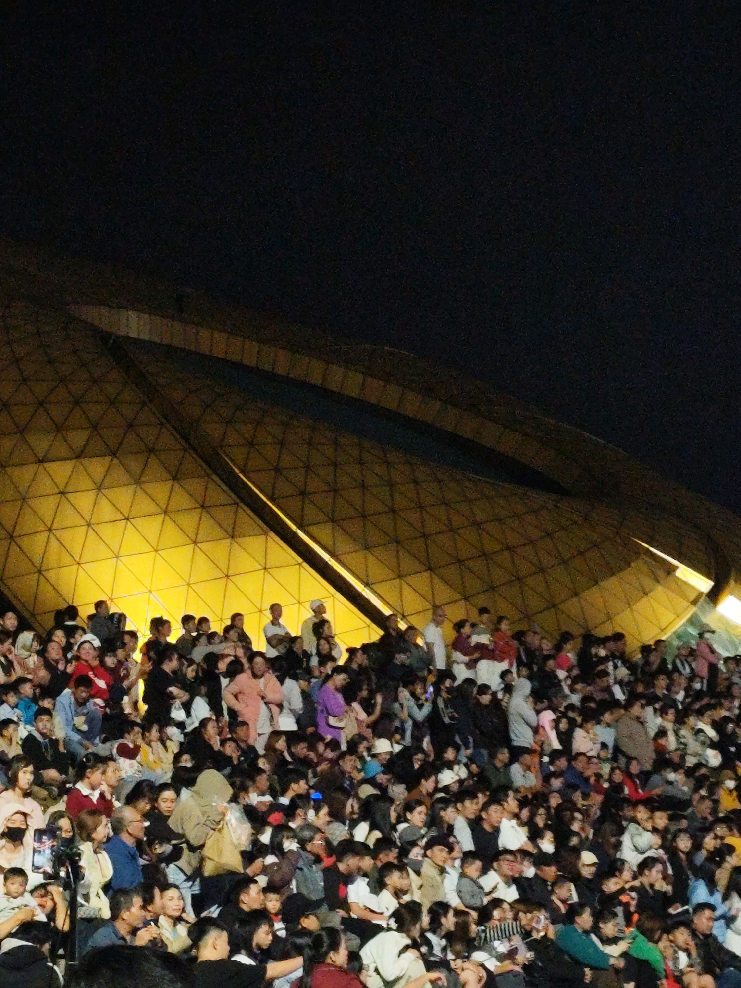 The image depicts a large crowd of people gathered outdoors at night, under a dimly lit, geometrically patterned golden structure. The structure, possibly a roof or canopy, has triangular facets and emits a warm yellow light, creating a contrast with the dark night sky. The crowd is densely packed, with individuals of various ages and ethnicities visible. Many are looking upwards and towards something out of frame, suggesting they are attending an event, performance, or viewing. The setting appears to be an open-air venue, potentially a plaza or amphitheater. The overall atmosphere is one of public gathering and shared experience under artificial illumination. While the image does not explicitly show the city of Da Lat, Vietnam, such architectural designs and public events are common in tourist destinations there.
