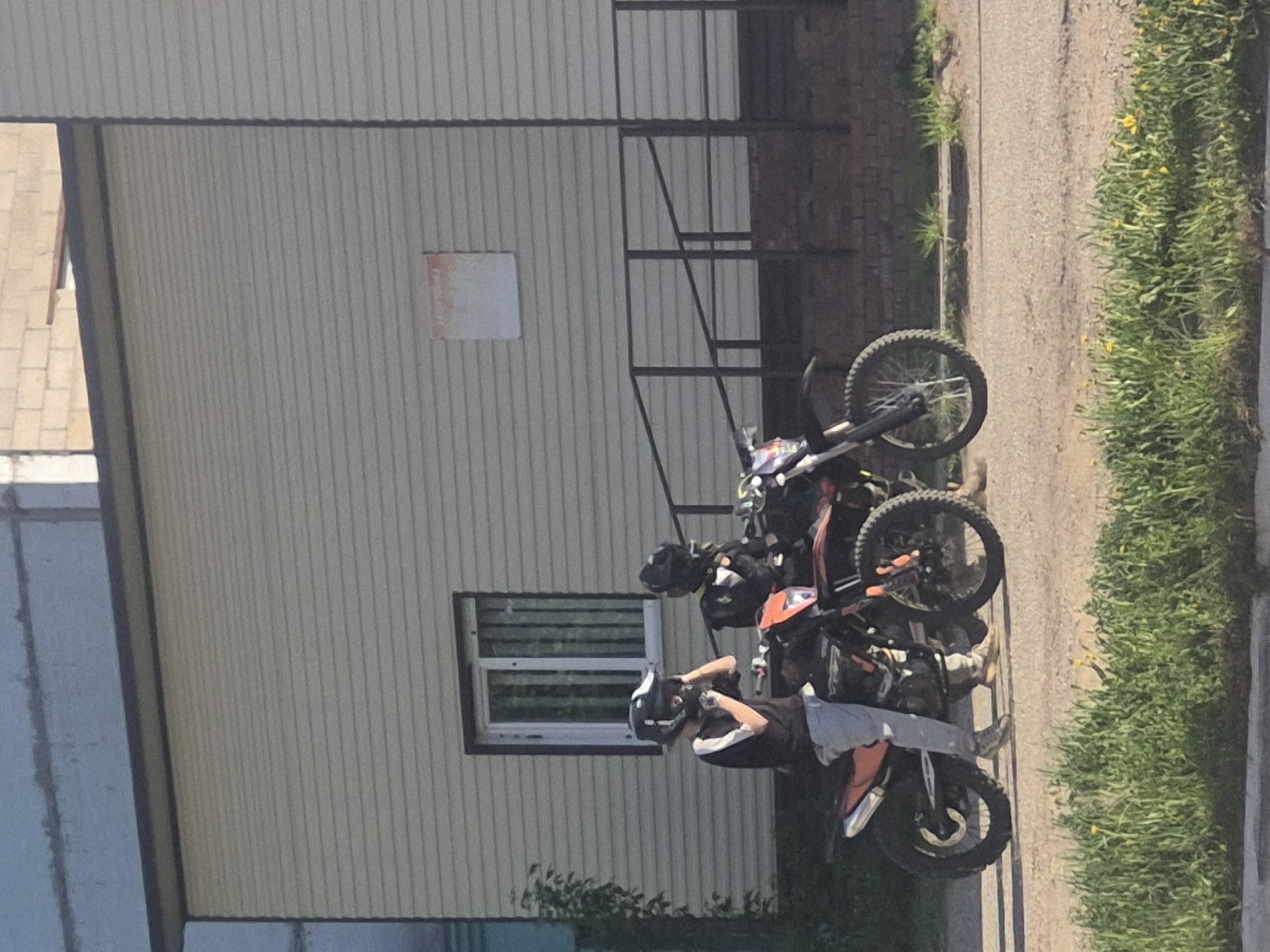 The image shows three parked dirt bikes next to a building on a street. A person wearing a helmet is standing next to the closest dirt bike. The building has a window and a metal ladder or railing is next to it. The street is lined with grass and weeds. The city and specific location are unknown.