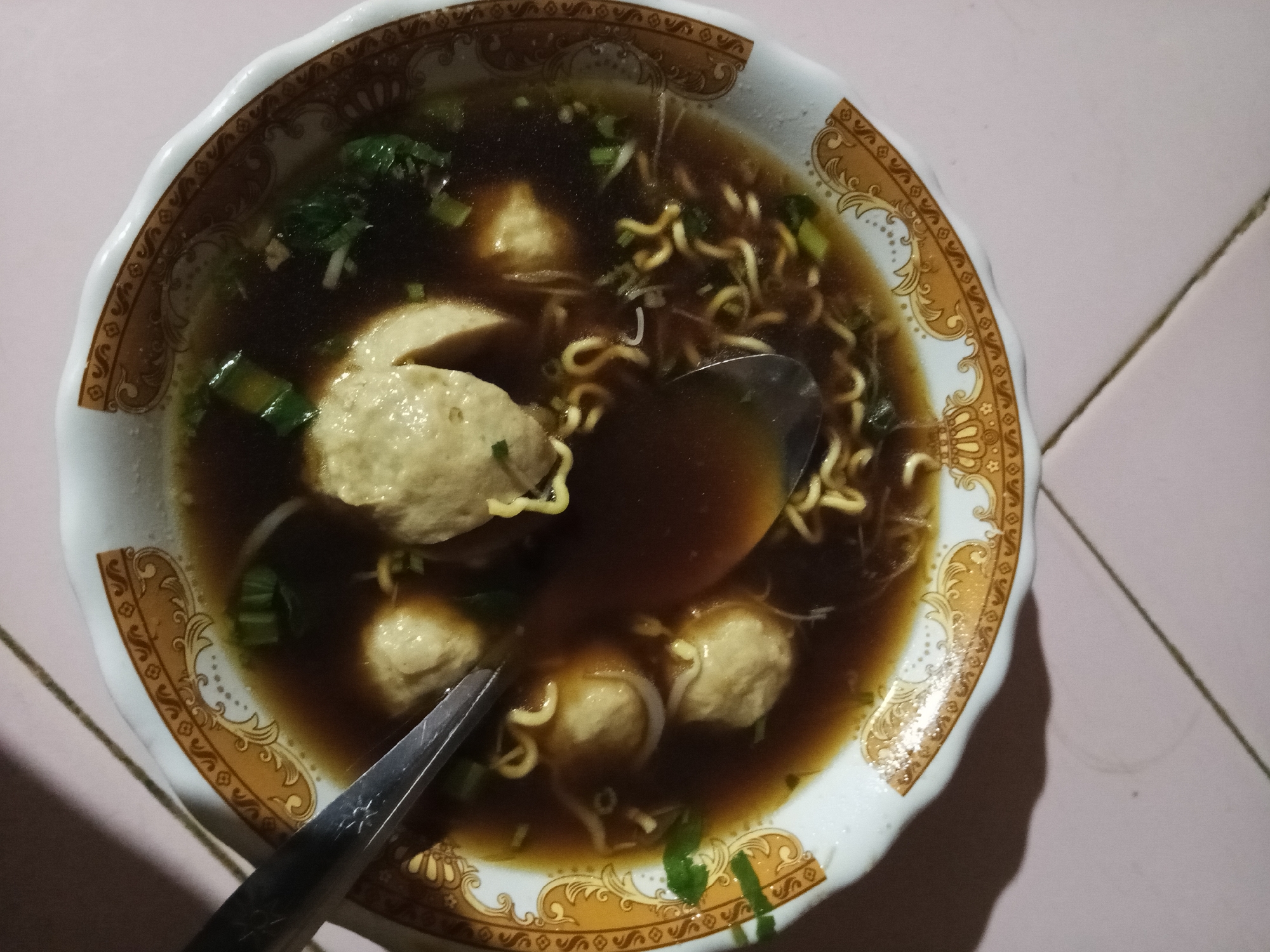 A close-up overhead shot shows a bowl of mie ayam, a popular Indonesian noodle dish, filled with dark broth, round meatballs, yellow noodles, and chopped green onions. Two spoons are submerged in the soup, with one angled towards the right and the other towards the left, suggesting the meal is being consumed. The bowl itself is white with a decorative golden trim and rests on a light pink tiled surface, possibly a table. The lighting suggests an indoor setting, likely at night. This dish is commonly found in local eateries and street food stalls throughout cities like Lubuklinggau, Indonesia.