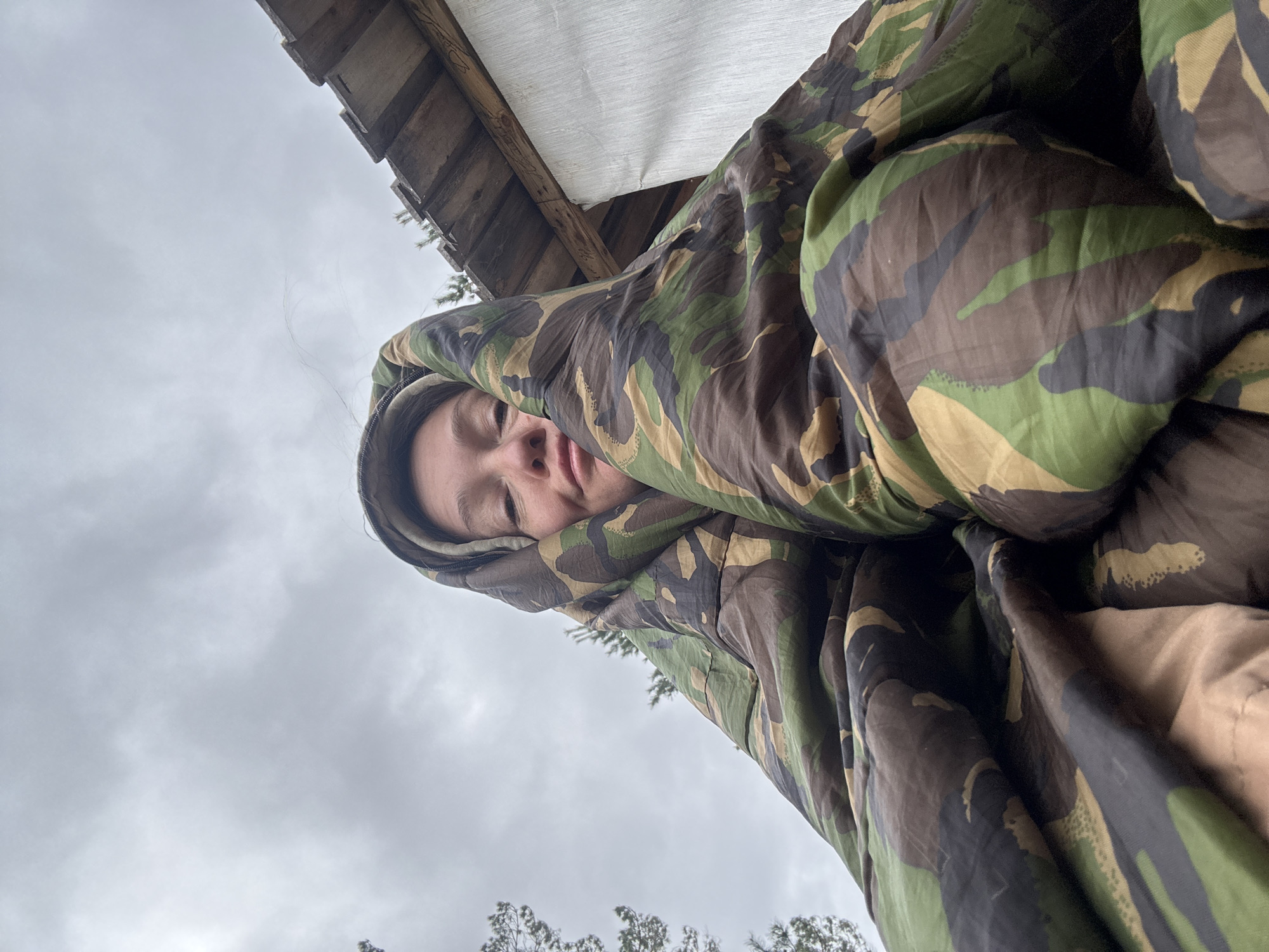 A person is enclosed in a camouflage sleeping bag, lying beneath a makeshift wooden shelter. The sky is overcast with gray clouds. Sparse trees are visible in the distance. The location is not determinable from the image.