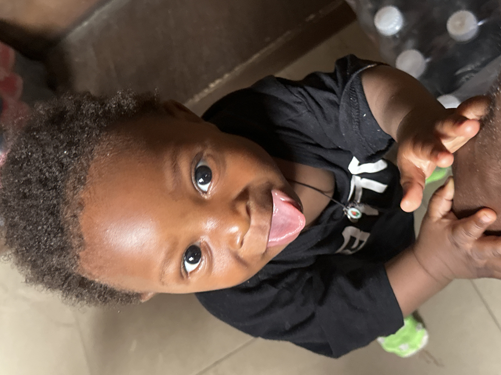 A high-angle photograph shows a baby with dark skin and dark, curly hair looking upwards directly at the camera. The baby has wide, dark eyes and a playful expression, with their tongue sticking out. They are wearing a black long-sleeved t-shirt with white letters partially visible on the front, possibly spelling "THE". A thin black necklace with a small, round pendant featuring red and green elements is worn around the baby's neck.
The baby's left hand is extended, appearing to grasp or touch the upper arm/shoulder of an adult, whose dark-skinned arm is partially visible on the right side of the frame. In the blurred background, several clear plastic bottles with white caps can be seen, likely water bottles, resting on a dark surface. The floor beneath the baby is a light-colored, possibly tiled, surface. The scene conveys a moment of playful interaction and curiosity, set indoors in what could be a home in Jos, Nigeria.