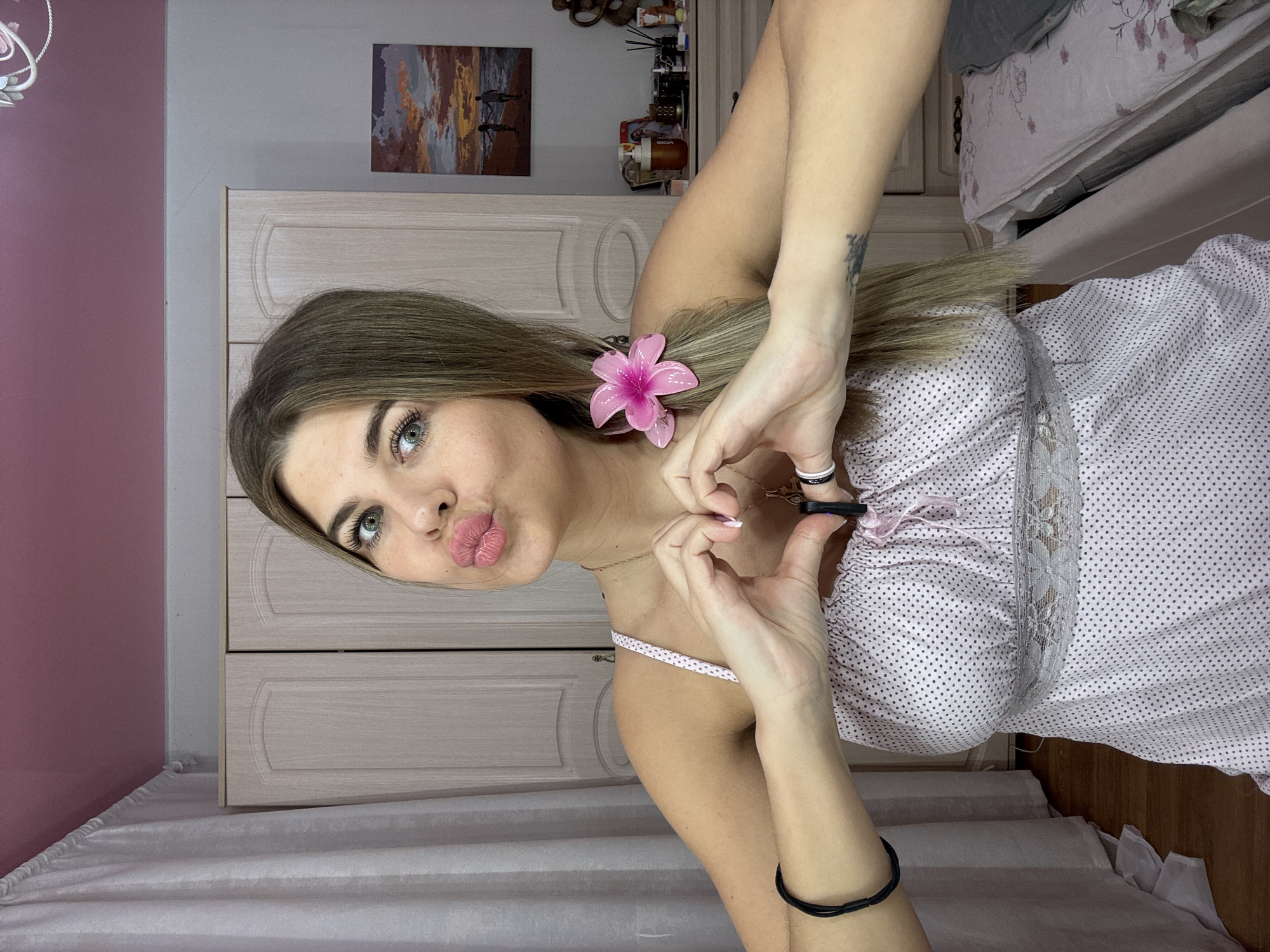 A young woman with shoulder-length blonde hair is posing indoors. She is wearing a light pink dress with white polka dots and lace detailing. A pink artificial flower is clipped into her hair. She is making a kissing face and forming a heart shape with her hands. She is wearing rings on her fingers. The background includes a light-colored wardrobe, a landscape painting, and personal items on top of the wardrobe. The location appears to be inside a residential building. There is no definitive information that indicates this image was taken in Chaikovsky, Russia.