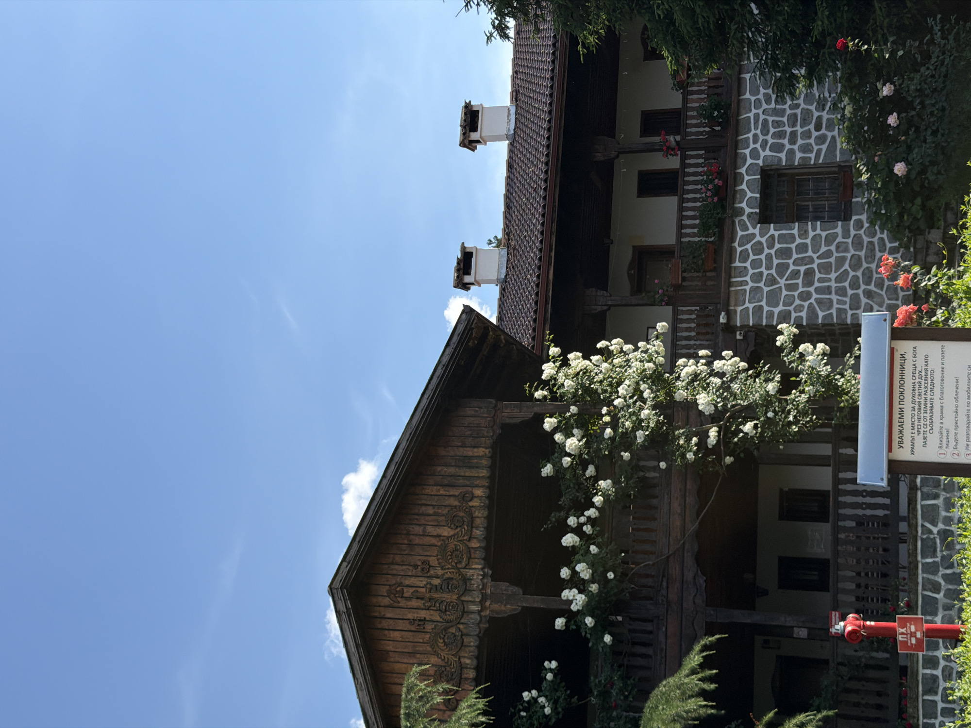 The image shows a building with a wooden upper story and a stone-clad lower story. There is a covered balcony on the upper level with a wooden railing, adorned with a large white climbing rose bush. The roof is tiled, and two white chimneys with dark caps rise from it. The building is set against a bright blue sky with scattered white clouds. In the foreground, there is a red fire hydrant with a sign, and a white information board with text in Bulgarian. Red and pink roses are blooming in front of the stone wall of the building. The overall scene depicts a historic building with lush vegetation in an outdoor setting. The text on the information board translates to "Dear Pilgrims. The monastery is a holy place with... God's... blessed... Respect the holy place... and its surroundings." It also lists rules such as "1. Bow to the monastery with reverence and humility!" and "2. Dress modestly!"
