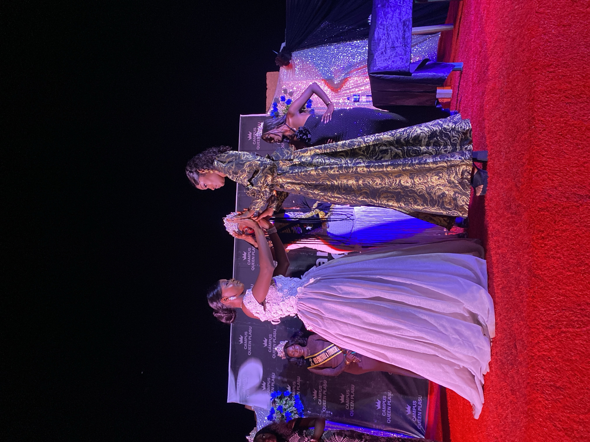 The image captures a pageant-style event, possibly in Durimi, Nigeria. Three women are the main subjects, presumably contestants or winners, wearing elaborate gowns and crowns. One woman wears a light, possibly white or silver, gown with floral details. Another woman is in a dark, possibly black or navy, gown with silver embellishments, positioned behind the others. The third is wearing a golden patterned gown with high neck and full sleeve.
The women are on a stage or platform covered in a red rug or carpet. The backdrop includes black fabric, shiny silver material, and a banner with logos reading "CAMPUS QUEEN PLASU." Lighting is dramatic with strong colors, possibly from stage lights. Other people are visible in the background.