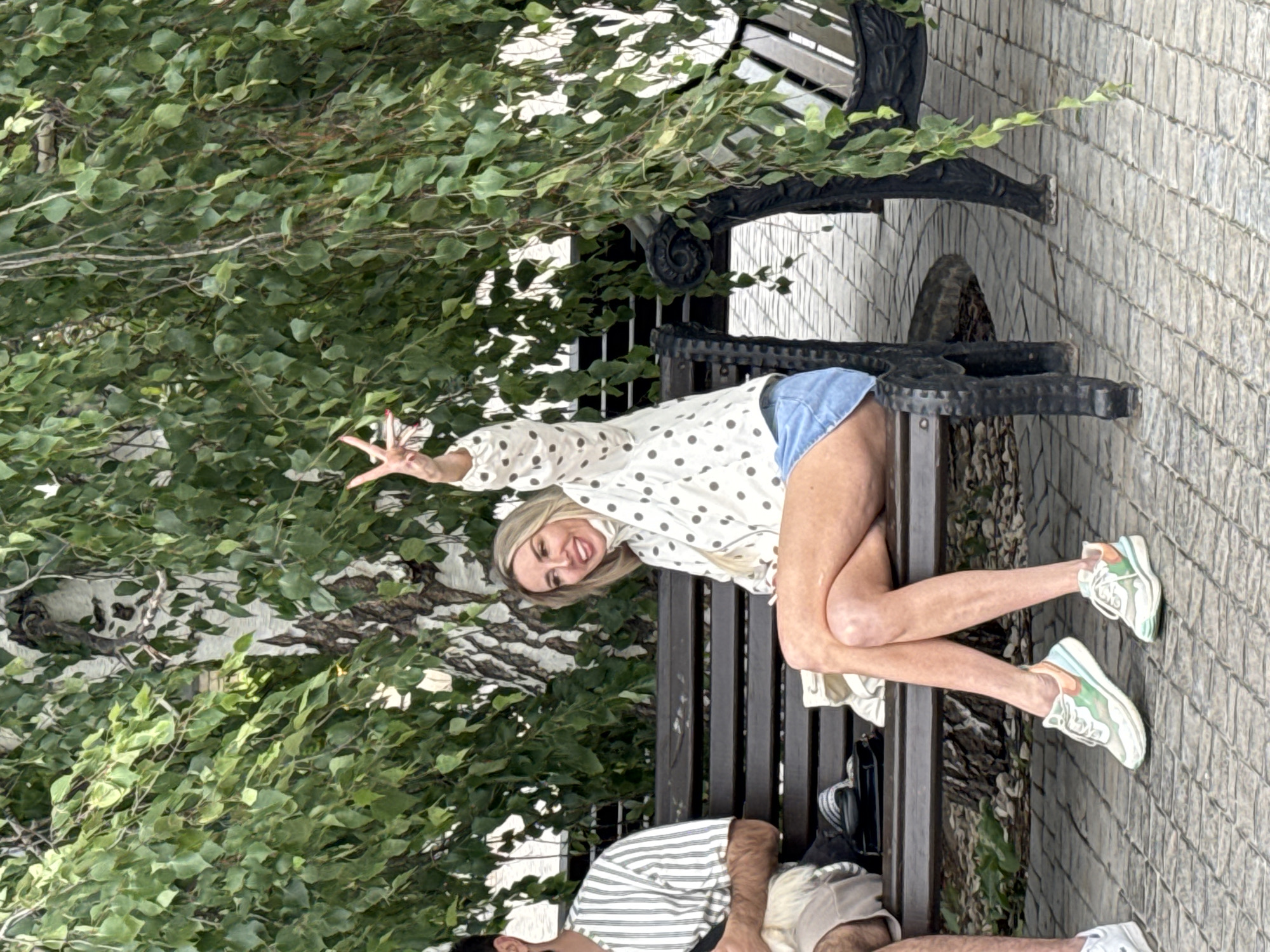 A medium shot captures a blonde woman sitting on a park bench in Khryashchevka, Russia, under the shade of a birch tree. She is wearing a white polka-dot jacket, a denim skirt, and colorful sneakers, and she is smiling and making a peace sign with her right hand. To her left, a man in a striped t-shirt is partially visible. The bench is made of dark wood and metal, and it is situated on a cobblestone path. Lush green foliage from the tree hangs over the scene, creating a dappled light effect.