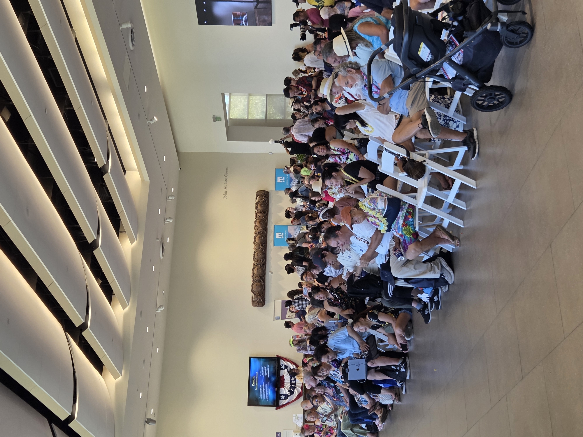 The image captures an indoor event in Santa Ana, California, within a large, brightly lit hall featuring a modern, slatted ceiling with integrated lighting. A diverse crowd of people, predominantly of Asian and Pacific Islander descent, is seated on white folding chairs, facing towards an unseen focal point. Many attendees are adorned with leis, suggesting a cultural or celebratory occasion. A large screen displays information, and various banners and logos are visible, including one for OCTA. A stroller is present in the foreground. The environment appears to be a public space, possibly a museum or community center.