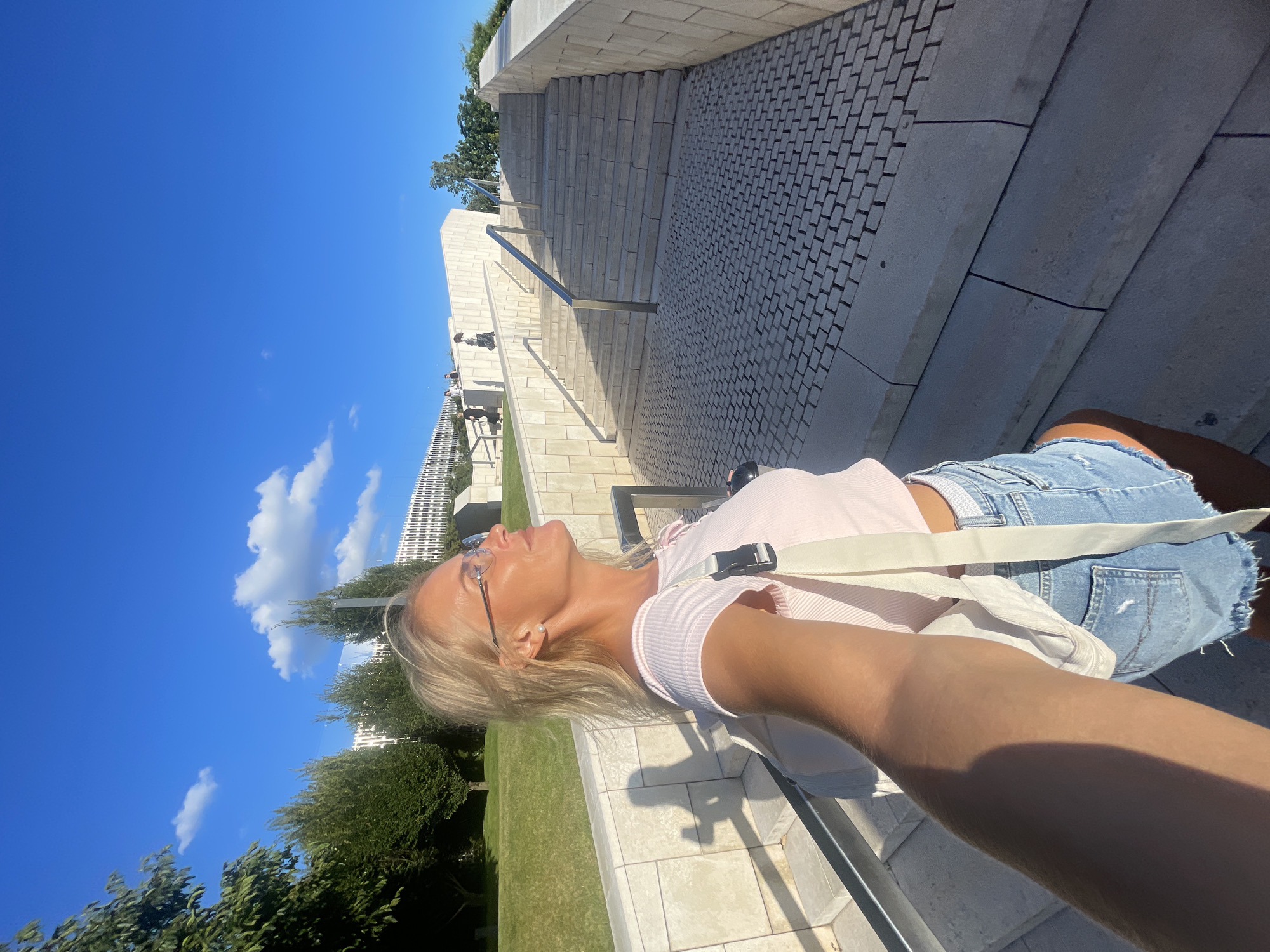 A young woman with blonde hair and glasses is taking a selfie outdoors on a sunny day. She is wearing a pink top and denim shorts. She is leaning back and looking up at the sky with her eyes closed, appearing relaxed and enjoying the sun. The background shows a modern architectural structure with stone steps and pathways, surrounded by lush green grass and trees. In the distance, a tall white building with many windows is visible. The sky is a clear, vibrant blue with a few scattered white clouds. The lighting suggests it is daytime, likely mid-morning or afternoon. The overall mood is serene and peaceful, capturing a moment of outdoor leisure. The scene appears to be a public space, possibly a park or an outdoor plaza in Krasnodar, Russia. There is no visible text in the image.