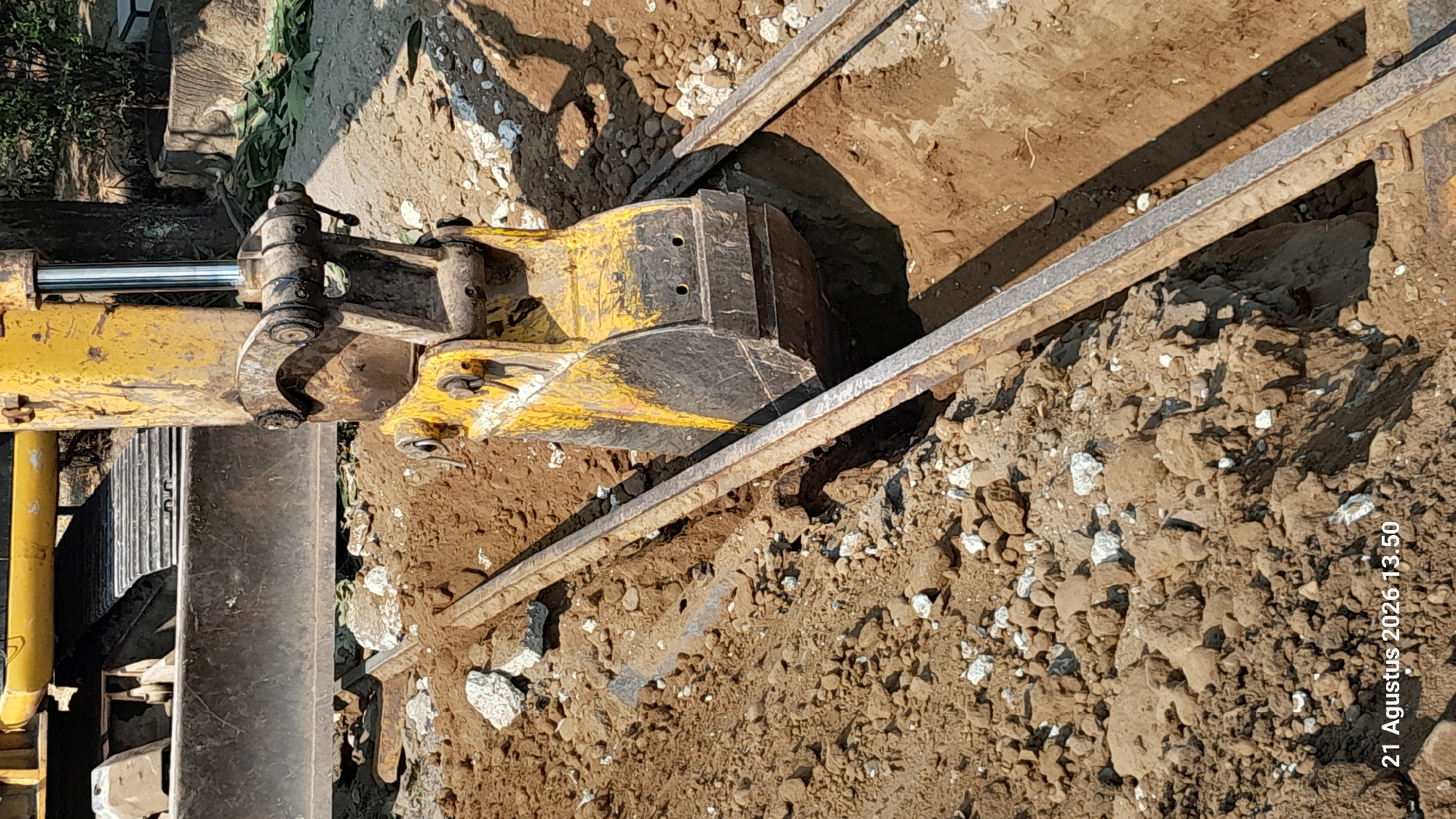 The image shows a close-up view of an excavator bucket digging a trench in the ground. The excavator is yellow and appears to be in active use, with its bucket partially submerged in the soil. Two parallel metal beams or rails are laid out along the trench, likely for guidance or support. The ground is composed of brown soil, rocks, and debris. The overall scene suggests ongoing construction or excavation work. A timestamp in the lower left corner reads "21 Agustus 2026 13.50".