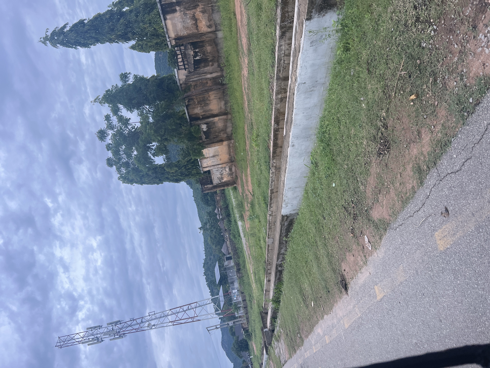 In the city of Kona Garu, Nigeria, a telecommunications tower stands tall on the left side of the frame, its red and white lattice structure reaching towards a cloudy sky. In the foreground, a paved road with yellow lane markings runs diagonally from the bottom left corner. Adjacent to the road is a grassy verge with a concrete drainage channel running alongside it. Beyond the drainage channel, a weathered, ochre-colored wall with occasional graffiti runs horizontally across the midground. Several tall, dark green trees stand behind the wall. In the background, a cluster of traditional buildings with thatched roofs are visible, nestled amongst lush green vegetation and rolling hills. The overall atmosphere is overcast and slightly humid. There are no people visible in the image.