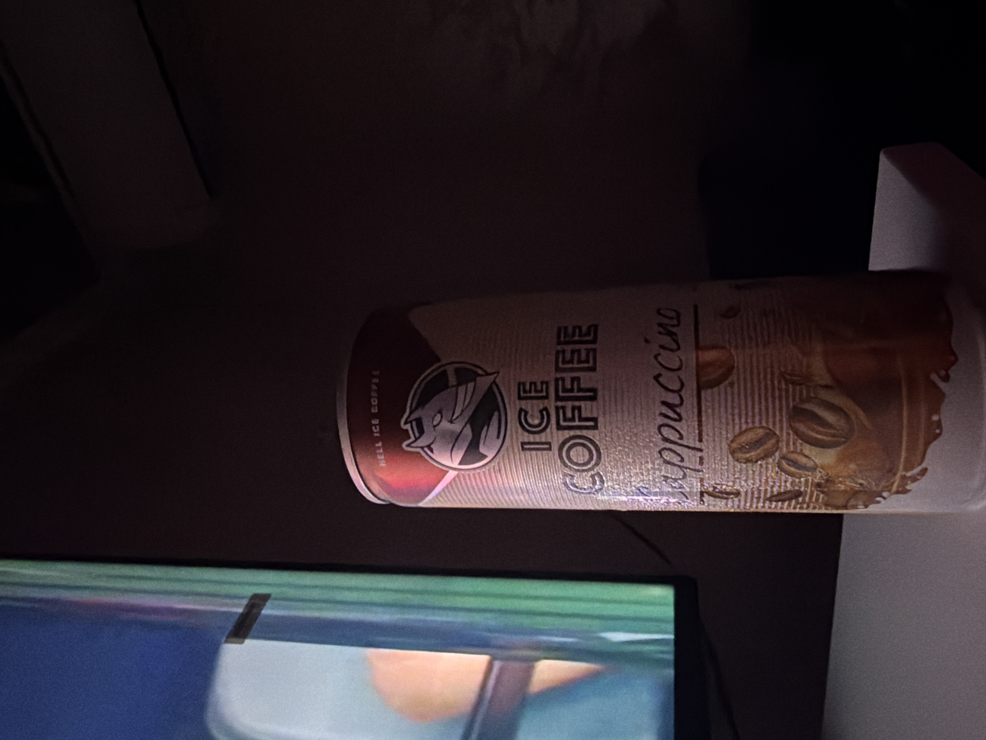 A close-up, low-angle shot shows a can of "ICE COFFEE" next to a television screen. The can, which has a picture of a devil-like character and coffee beans, is the primary subject. The television screen displays a blurry image of a person's arm. The scene is dimly lit, suggesting it is indoors, likely in Ognianovo, Bulgaria.