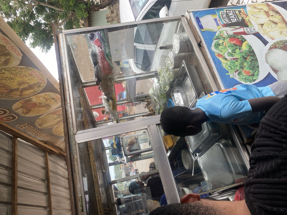The image shows a food stand with a glass front in Adenta Municipality, Ghana. The stand is made of metal and has a tin roof. It is located in a bustling street with traffic. On the roof of the stand is a sign advertising "Jain Rice" and fried chicken. The stand has a variety of food items for sale, including salad, fried chicken, and rice. A young boy in a blue shirt is standing in front of the stand looking at the food. Another adult, partially obscured, is standing to the left, looking at the boy and possibly at the food. Both are standing at the stand, waiting to be served. The stand is lit up with bright lights, making it an attractive and inviting place to buy food. The street outside the stand is busy with traffic and pedestrians.