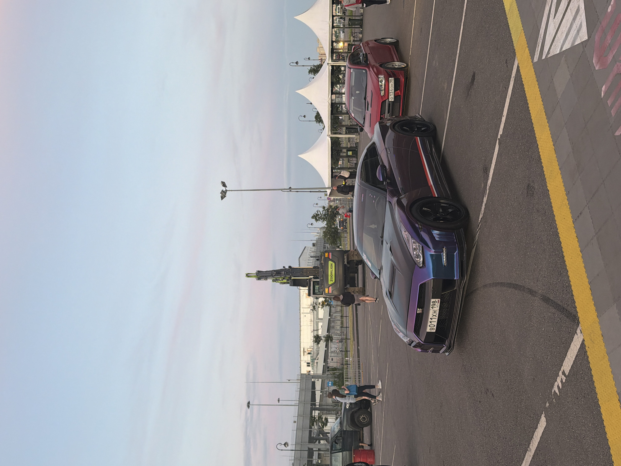 The image shows a parking lot at an outdoor event, possibly a car show, with several vehicles and people present. In the foreground, a purple Nissan GT-R is parked, displaying tire marks on the asphalt. Behind it, a red Mitsubishi Lancer Evolution is parked. Further in the background, a dark-colored SUV is visible, and a large construction excavator with "ZOMLION" branding is also present. Several individuals are walking around the vehicles and in the vicinity of white tents that appear to be part of the event setup. The sky is partly cloudy with soft pastel hues, suggesting it might be dusk or dawn. The overall environment appears to be an open, paved area with streetlights and some urban infrastructure visible in the distance.