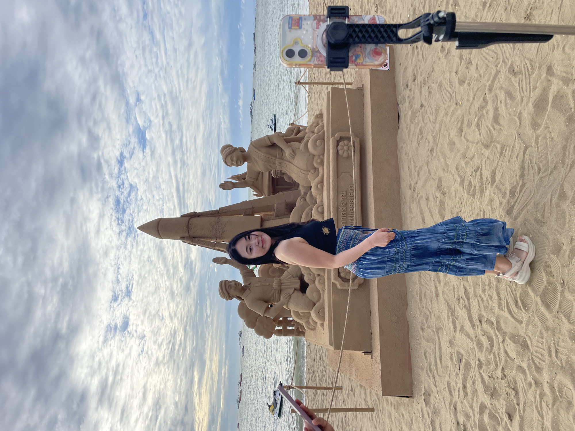 A young Asian woman stands on a sandy beach in Pattaya City, Thailand, posing in front of a large sand sculpture. The sand sculpture depicts a rocket-like structure flanked by two figures, a man and a woman, both with hands raised. The woman is wearing a black crop top and a blue tiered skirt, with white platform sandals. She is looking at the camera and smiling. In the background, the ocean is visible with several boats and jet skis. A phone on a selfie stick is set up to record the scene. The sky is overcast with scattered clouds. The sand sculpture is on a raised platform with some Thai writing.