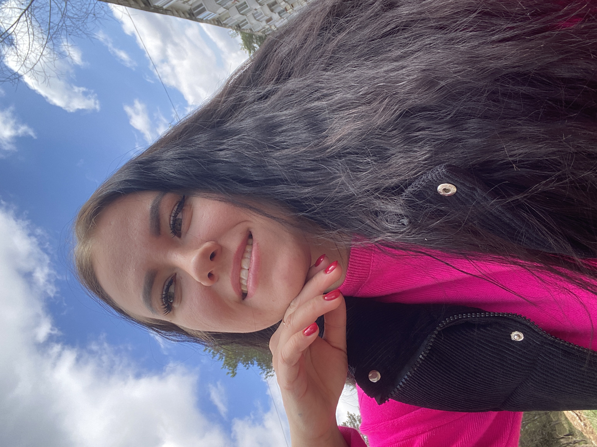 A woman with long dark hair is smiling at the camera. She is wearing a bright pink sweater and a black jacket. Her left hand is near her face. The background shows a blue sky with white clouds and a partial view of a building. The location is Chaikovsky, Russia.