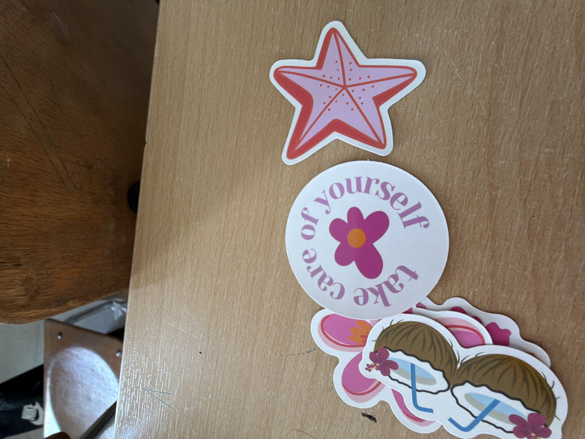 The image shows a close-up of a wooden surface with several stickers. In the lower left quadrant, there are a cluster of stickers featuring floral designs and a pair of sunglasses made from coconuts. To the right of these, a circular sticker with pink text that reads "take care of yourself" surrounds a pink flower with an orange center. Further to the right, a pink and red starfish sticker is visible. The background is blurred and appears to be part of a classroom or office setting with a wooden chair and a white surface. There is no indication of the location being in Burgas, Bulgaria.