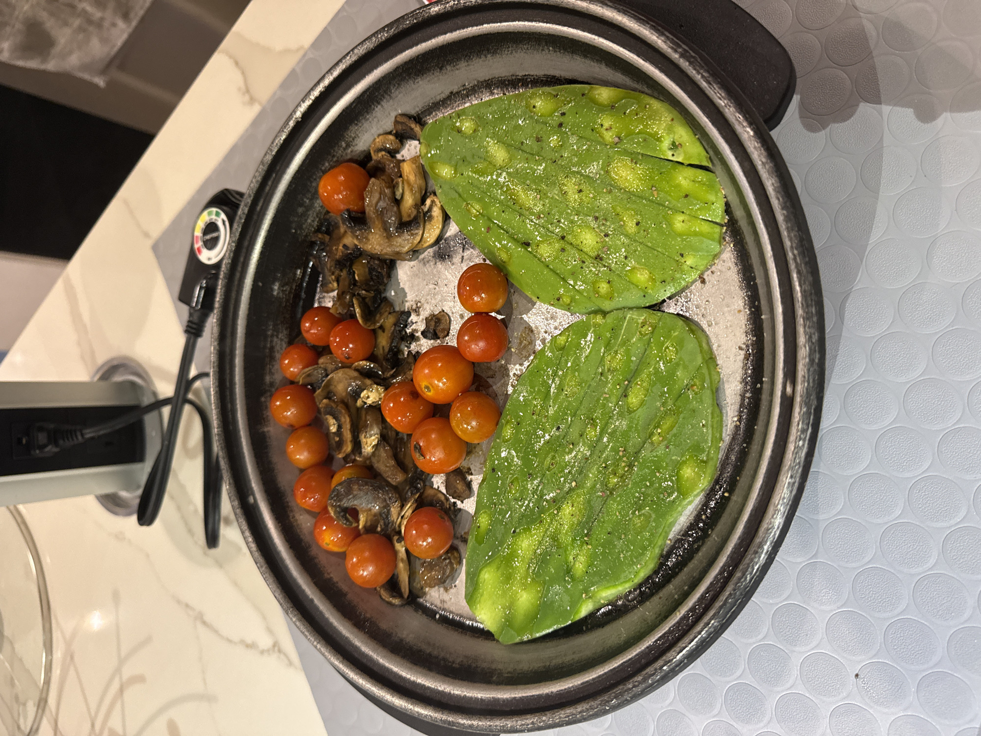 The image shows a stainless steel pan containing two green nopal cactus pads seasoned with pepper and cooked next to sauteed mushrooms and cherry tomatoes. The pan is on a flat surface with a power cord and temperature control knob extending off the left side. The surface below the pan is either a countertop or tabletop, and the background includes partial views of a countertop, cabinet, and a patterned surface. The location and specific setting details for San Bernardino Tlaxcalancingo, Mexico, are not determinable from the image content.