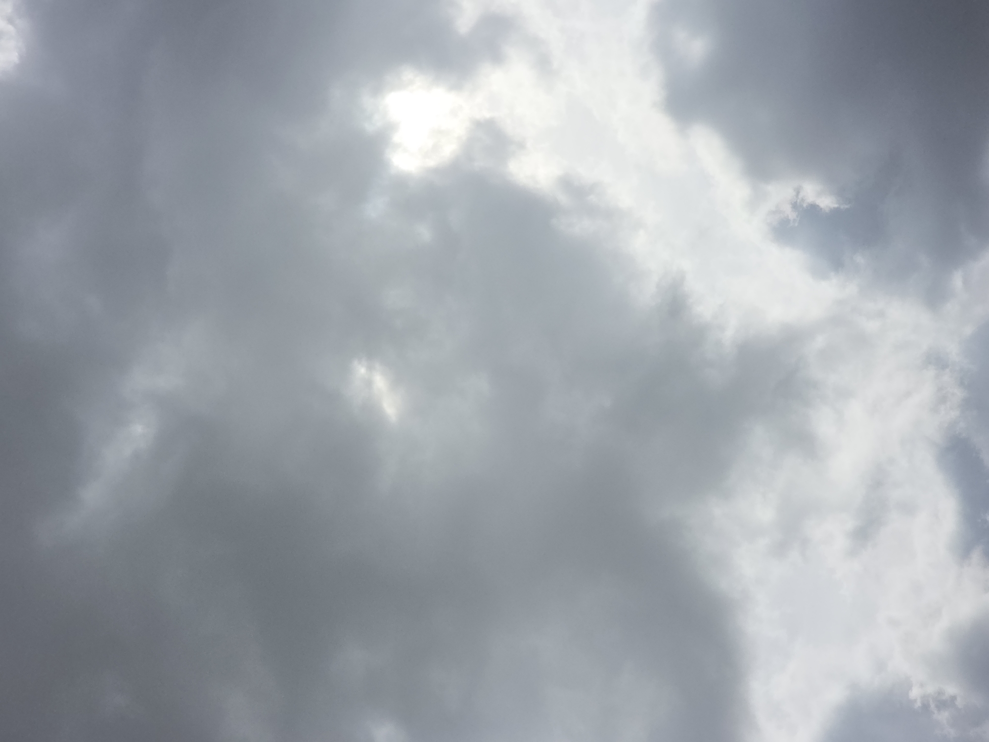 The image shows a cloudy sky over Qarabalıq, Kazakhstan. The clouds are a mix of dark gray and lighter, almost white formations, suggesting a partly overcast or stormy day. Sunlight is breaking through some of the clouds, creating bright patches and illuminating the sky from behind the denser cloud cover. There are no visible structures, people, or animals in the frame, making it a purely atmospheric scene. The focus is solely on the dynamic texture and light variations within the cloudscape.