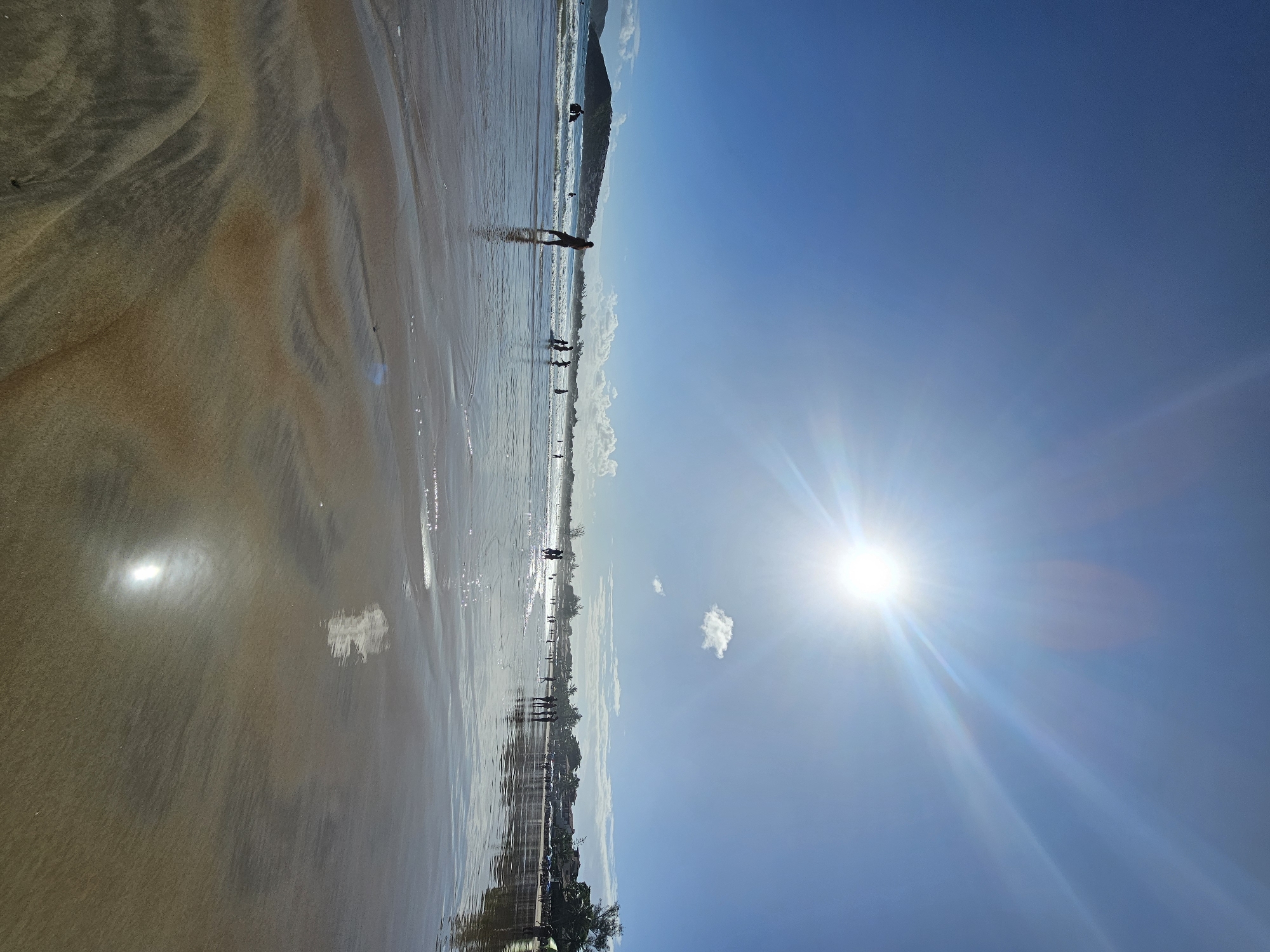 The image depicts a beach scene in Armação dos Búzios, Brazil, with a clear blue sky and bright sunlight. The wet sand reflects the sky and sunlight, creating a mirror-like effect. Several people are visible along the shoreline, some standing and some walking. The beach is flat and wide, with gentle waves lapping at the shore. The background features a low-lying landmass. A small cloud is in the sky to the left of the sun. The angle of the photo implies that it was taken facing east.