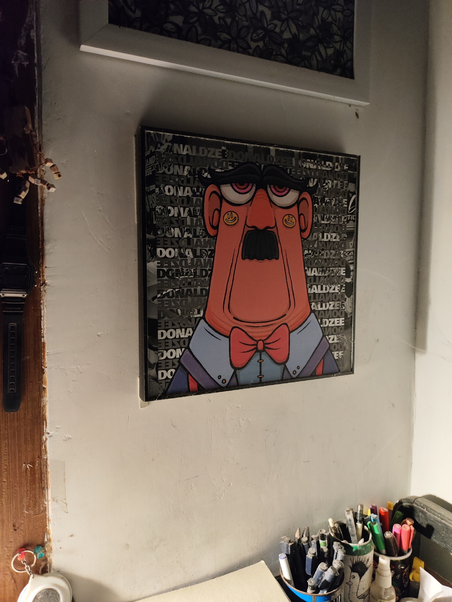 The image depicts a framed artwork hanging on a textured, off-white wall. The artwork features a stylized cartoon character with a large, round, reddish-orange face, a prominent black mustache, pink eyes with yellow smiley faces as pupils, and a red bowtie. The background of the artwork is a collage of black and white text, some of which appears to be repeated words like "DONALDZEE." To the left of the artwork, a black digital watch is attached to a wooden surface that appears to be a door frame or furniture. Below the watch, a white keychain with colorful beads is visible. In the lower right of the image, a collection of pens and markers in various containers is arranged on a surface, with some of the pens being actively used or sorted. The scene appears to be an indoor space, possibly a studio or personal workspace, in the city of Kepung, Indonesia.