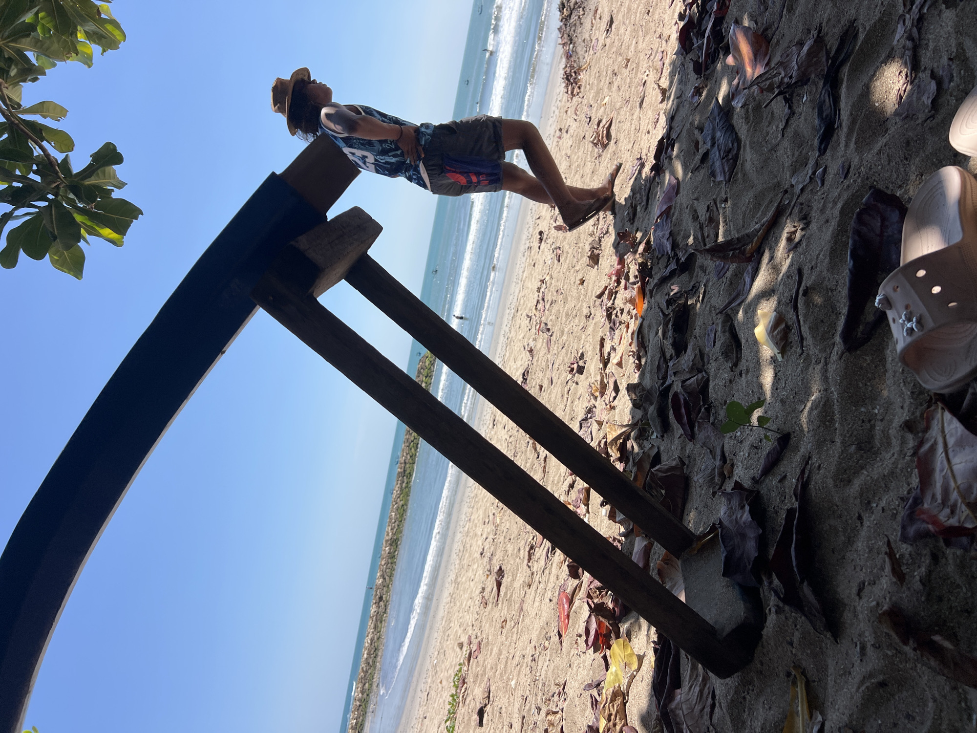 A man in a tropical hat and a tank top walks on a sandy beach, with the ocean and a breakwater in the background. He is leaning against a large, wooden structure that is partially buried in the sand. The structure appears to be a part of an abandoned or in-progress construction project, possibly a stage or a shelter. Fallen leaves and debris are scattered on the sand, suggesting a natural, unkempt environment. The sky is clear blue, indicating a sunny day. The scene is set in Kuta, Indonesia, a popular tourist destination known for its beaches and surf.