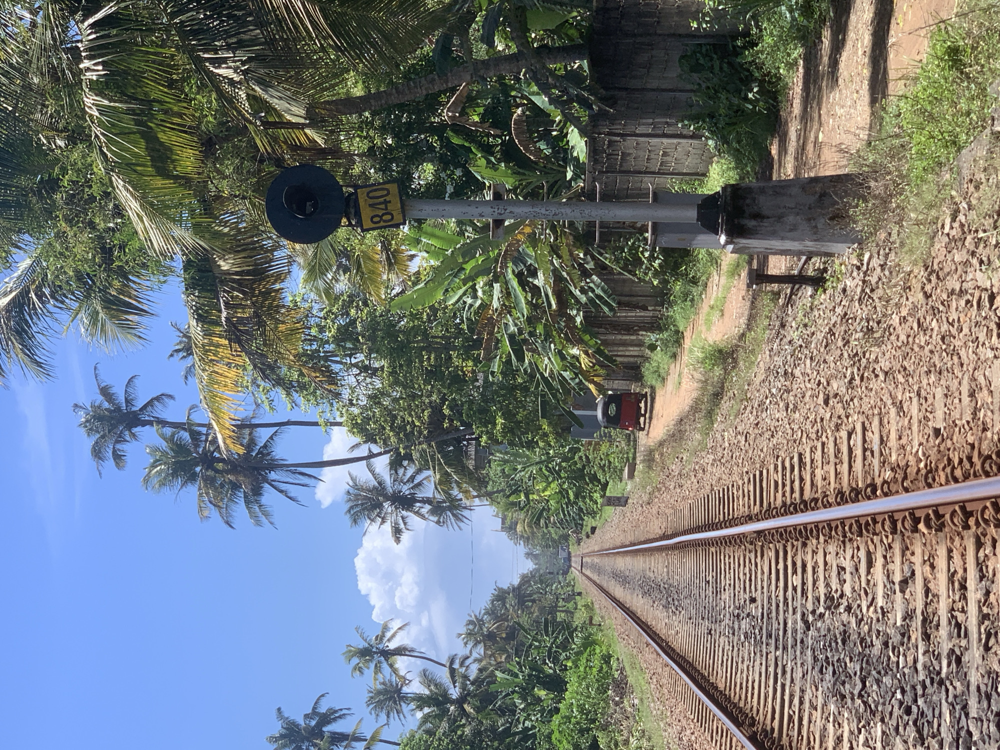 In Ahangama, Sri Lanka, a railway track stretches into the distance, flanked by lush tropical vegetation and palm trees. A red and black tuk-tuk is parked on a dirt road to the right of the tracks, near a signal post displaying the number 840. The sky is bright blue with scattered white clouds. The scene depicts a typical rural landscape of the region.