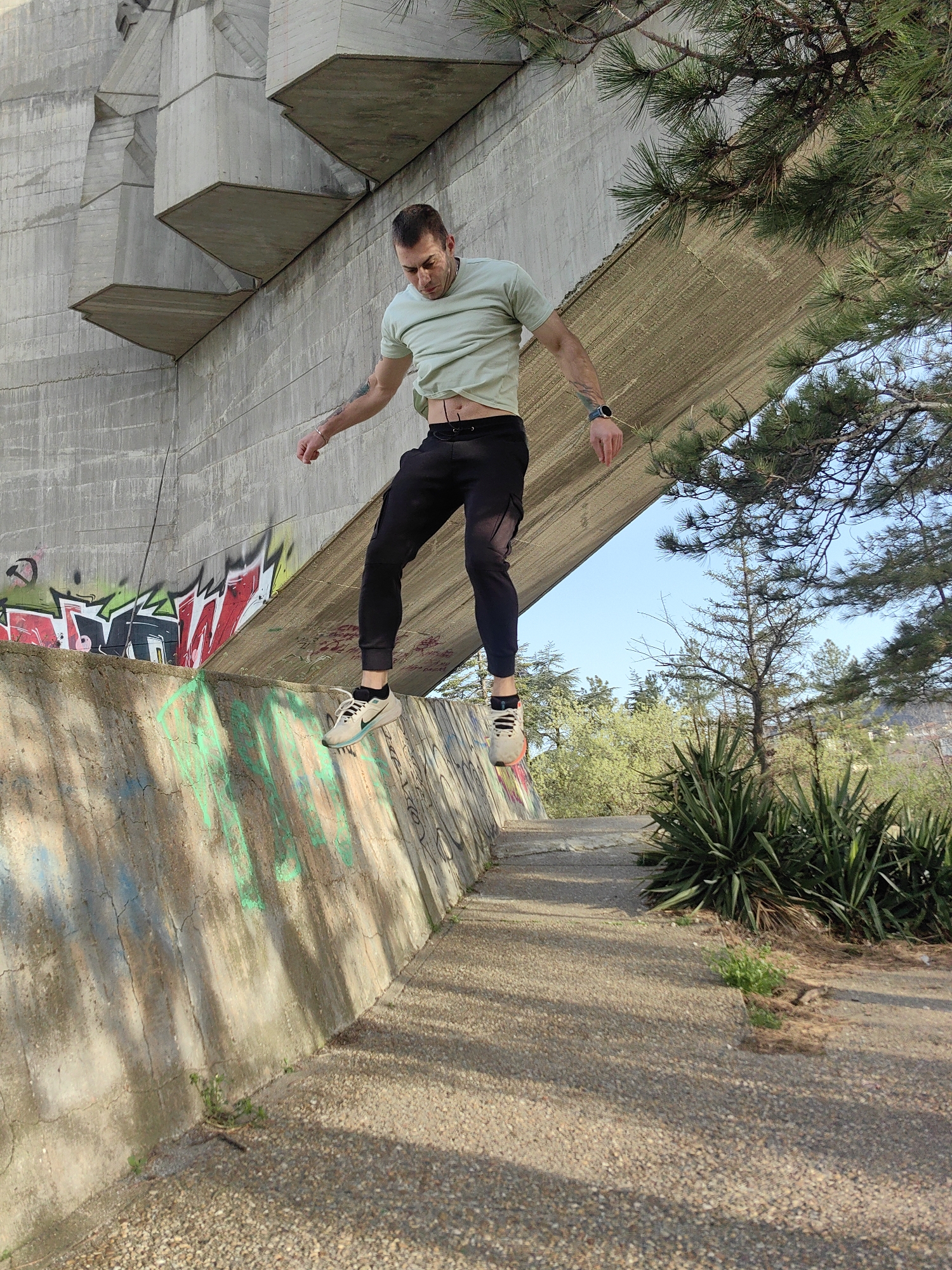 A man is jumping down from a concrete ledge in a park-like area. He is wearing a light green t-shirt, black pants, and athletic shoes. His arms are slightly outstretched for balance, and he is looking down. The ledge is graffiti-covered and bordered by a sidewalk or path made of small stones.
Overhead, a large concrete structure, possibly a bridge or an element of architecture, angles down towards the ground, creating a sense of enclosure. The structure has geometric, staggered patterns on its side. Part of the structure appears to be a component of the Asparuhov Bridge in Varna, Bulgaria. Trees and shrubbery surround the location, suggesting an outdoor, possibly urban, setting. The sky is partially visible and light blue.