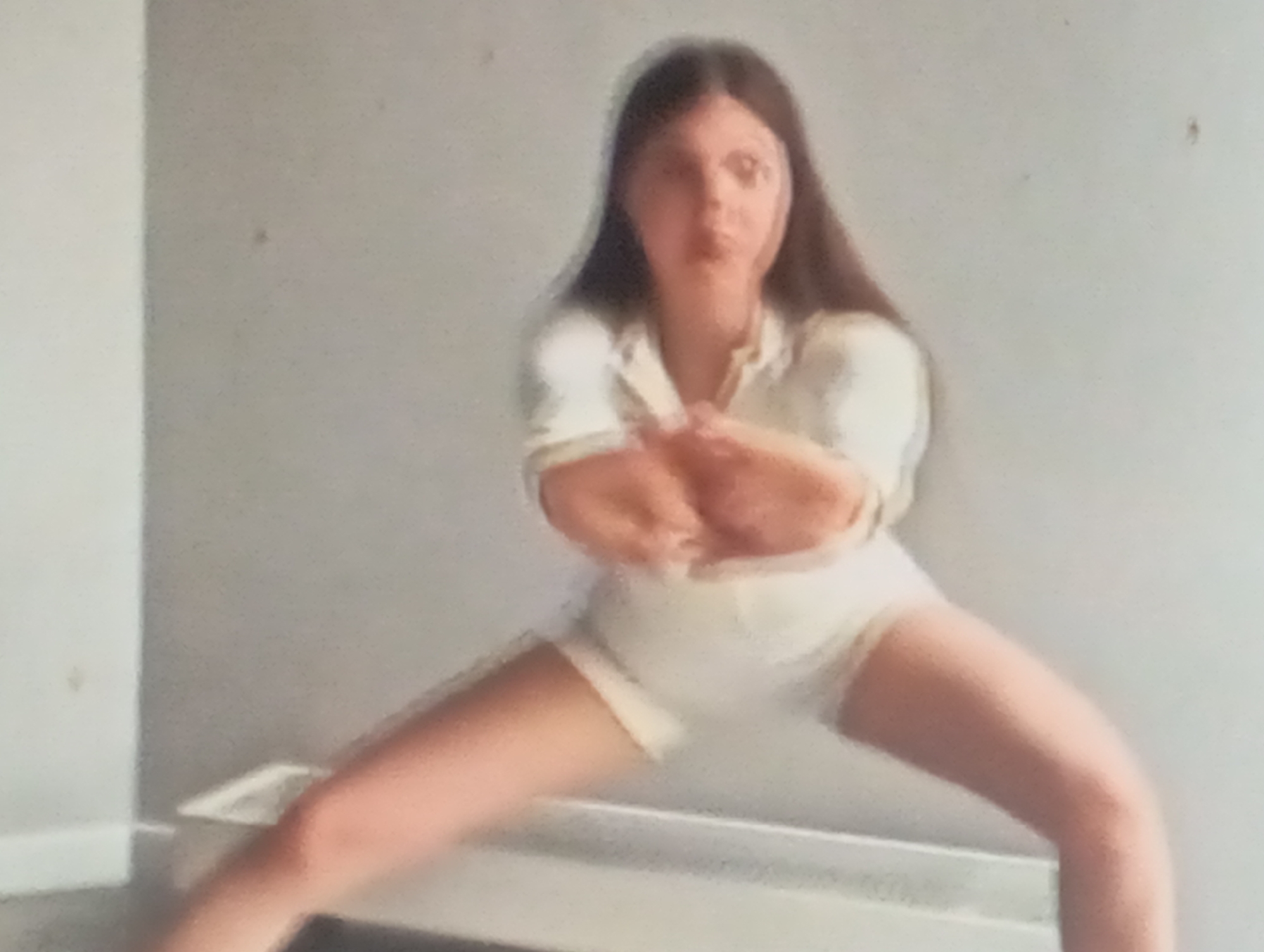 In Sofiivska Borshchahivka, Ukraine, a person with long, dark hair is captured in a low-angle shot. They are wearing a white collared shirt and white shorts. The individual is in a wide-legged squatting position, with their arms crossed in front of their chest. The background consists of a plain, light-colored wall and a white baseboard.