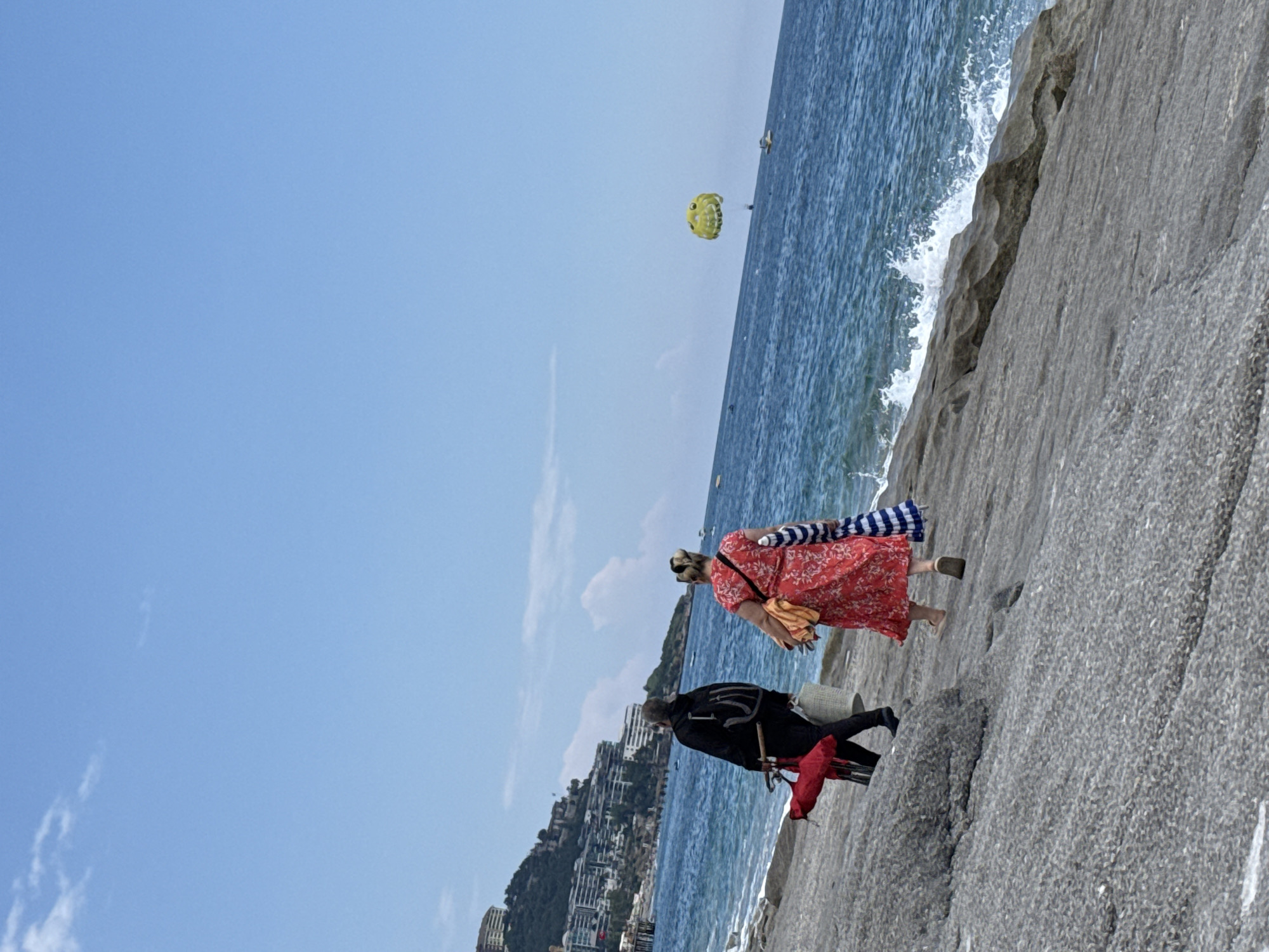 Captured in the city of Unknown, Unknown, the photograph depicts a coastal scene. Two individuals are walking along a stone embankment by the sea. The first person, clad in a black jacket and trousers, is carrying a red object. The second person, dressed in a red floral dress, has a bag slung over their shoulder and carries a striped blanket. The sea is visible on the right side of the image, with a yellow parasail present in the sky. In the background, a hill with buildings is seen. The sky is predominantly blue.