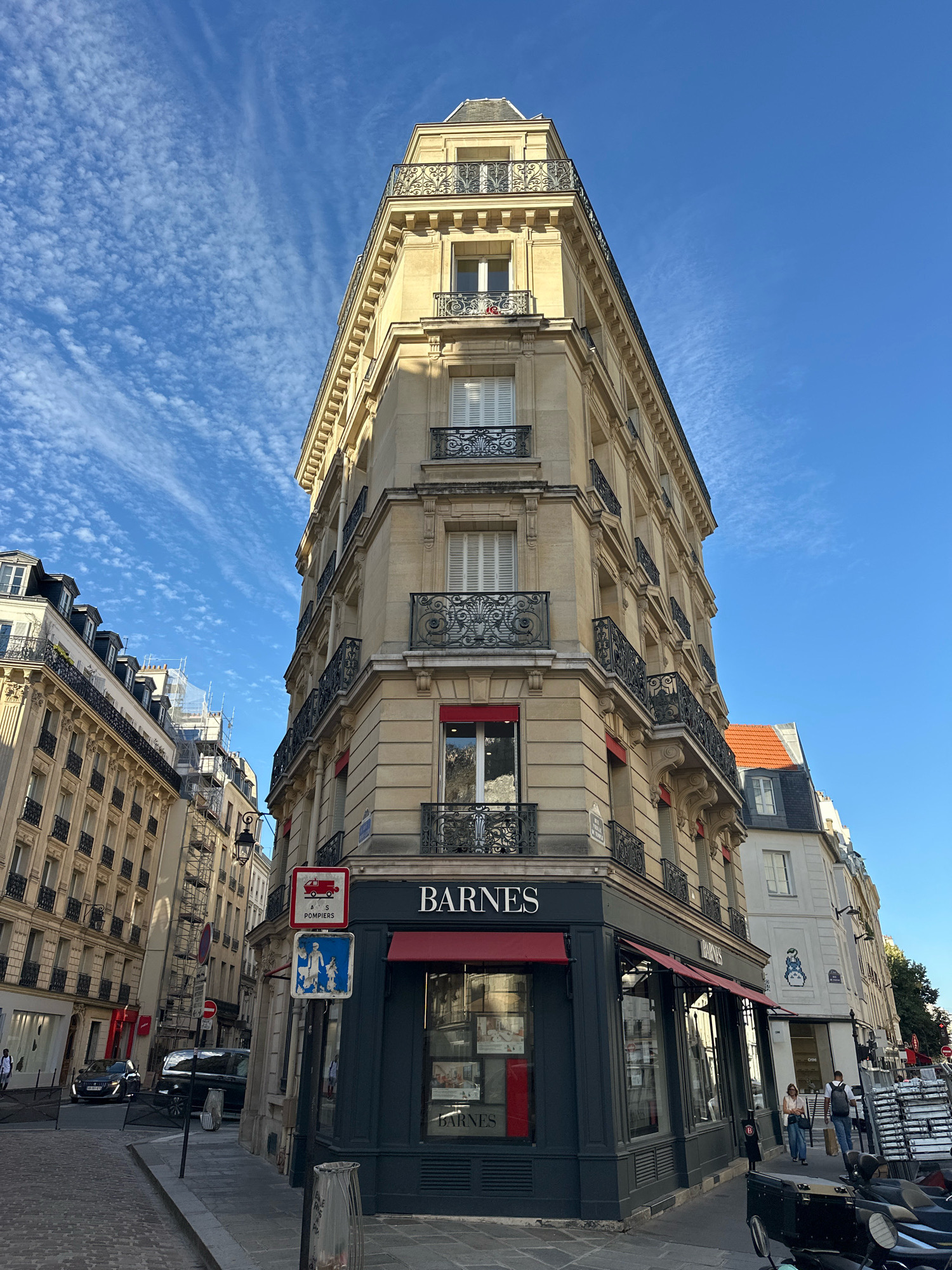 The image shows a corner building in Paris, France, with a Barnes real estate agency on the ground floor. The building is tall and has a distinctive triangular shape, with multiple floors featuring ornate balconies and windows. The sky is a clear blue with scattered wispy clouds.
On the sidewalk in front of the Barnes agency, there are several street signs, including one with a red fire truck symbol and "POMPIERS" written below it, indicating a fire department related sign. Another sign depicts stick figures of people. A few people are walking on the sidewalk, one of whom is carrying shopping bags. Several cars are visible on the street, including a black SUV and a darker vehicle parked further down. The street is paved with cobblestones. Adjacent buildings of similar architectural style line the street. There are also some motorcycles parked on the side of the street.
