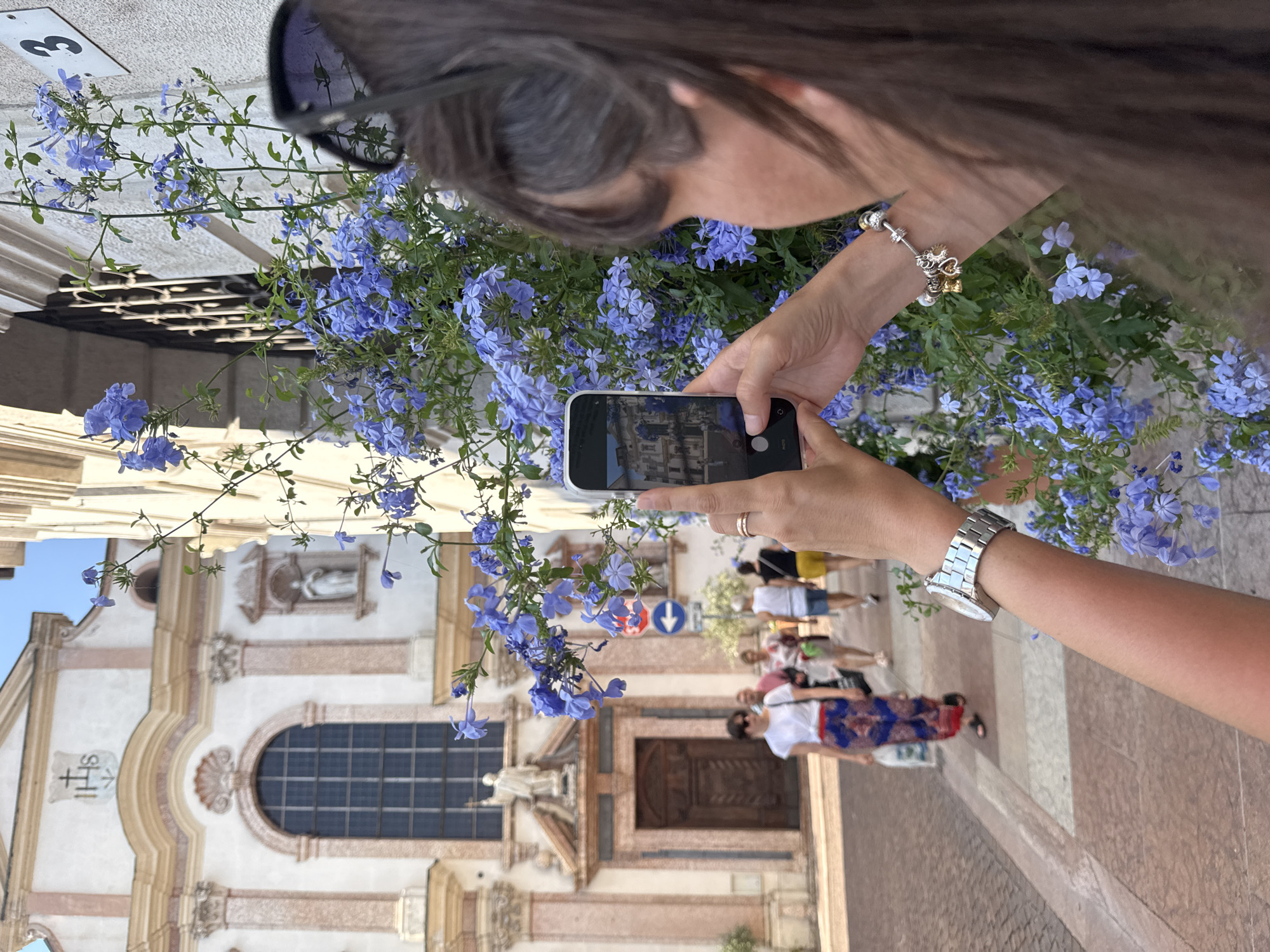 The image shows a person taking a photograph of a building with a smartphone. The building appears to be a church or a historic structure, with ornate architecture, a large arched window, and religious iconography. The person is holding the phone in landscape orientation, and the screen displays a preview of the photo being taken, showing the building. The person is wearing sunglasses on their head and a bracelet on their wrist. The foreground is filled with blooming blue flowers, which are also in focus in the lower part of the smartphone screen. In the background, other people are walking on a paved street. The scene is outdoors, suggesting a daytime setting. The weather appears to be clear and sunny. The overall mood is one of leisurely sightseeing and capturing memories. The image appears to be taken in Trento, Italy, given the visual cues of the architecture.
