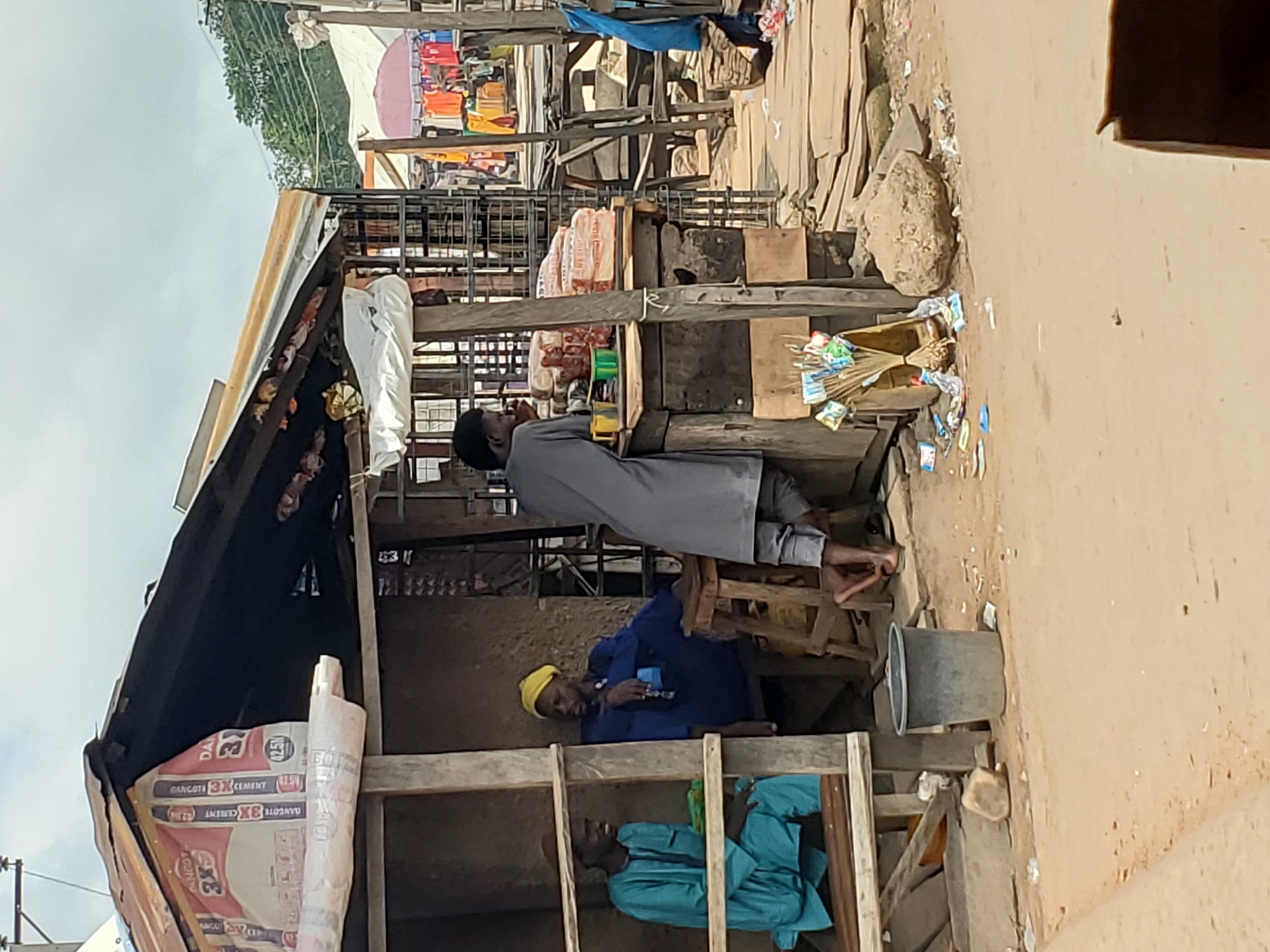 The image depicts a bustling street market in Mararaba, Nigeria, under a bright daytime sky. The scene is characterized by informal, makeshift stalls constructed primarily from rough wooden planks and corrugated iron sheets, set on an unpaved, dusty ground scattered with debris.
Several individuals are visible within the market environment. In the mid-ground, a man in a grey long-sleeved garment stands upright, leaning against a wooden support structure within a stall. He appears to be barefoot or wearing very light footwear. Nearby, within another section of the wooden stall, a man wearing a blue shirt and a yellow cap is seated on a low wooden chair, looking down. Below him, another man dressed in a light blue traditional outfit is also seated, partially obscured by the wooden framework. In the background, another individual in a light-colored robe is partially visible near a stall.
The stalls display various goods for sale. Prominently featured are multiple bags of "DANGOTE 3X CEMENT (42.5R)," indicating the presence of a building materials vendor. A wooden box on the ground holds several brooms along with small packaged items, possibly snacks or household essentials. Other small items hang from the underside of a corrugated roof. A grey bucket sits on the ground near the seated individuals. In the background, beyond the immediate stalls, more market activity is suggested by visible clothes on display and colorful umbrellas. The overall impression is that of a lively, functional community marketplace with everyday commerce taking place.
