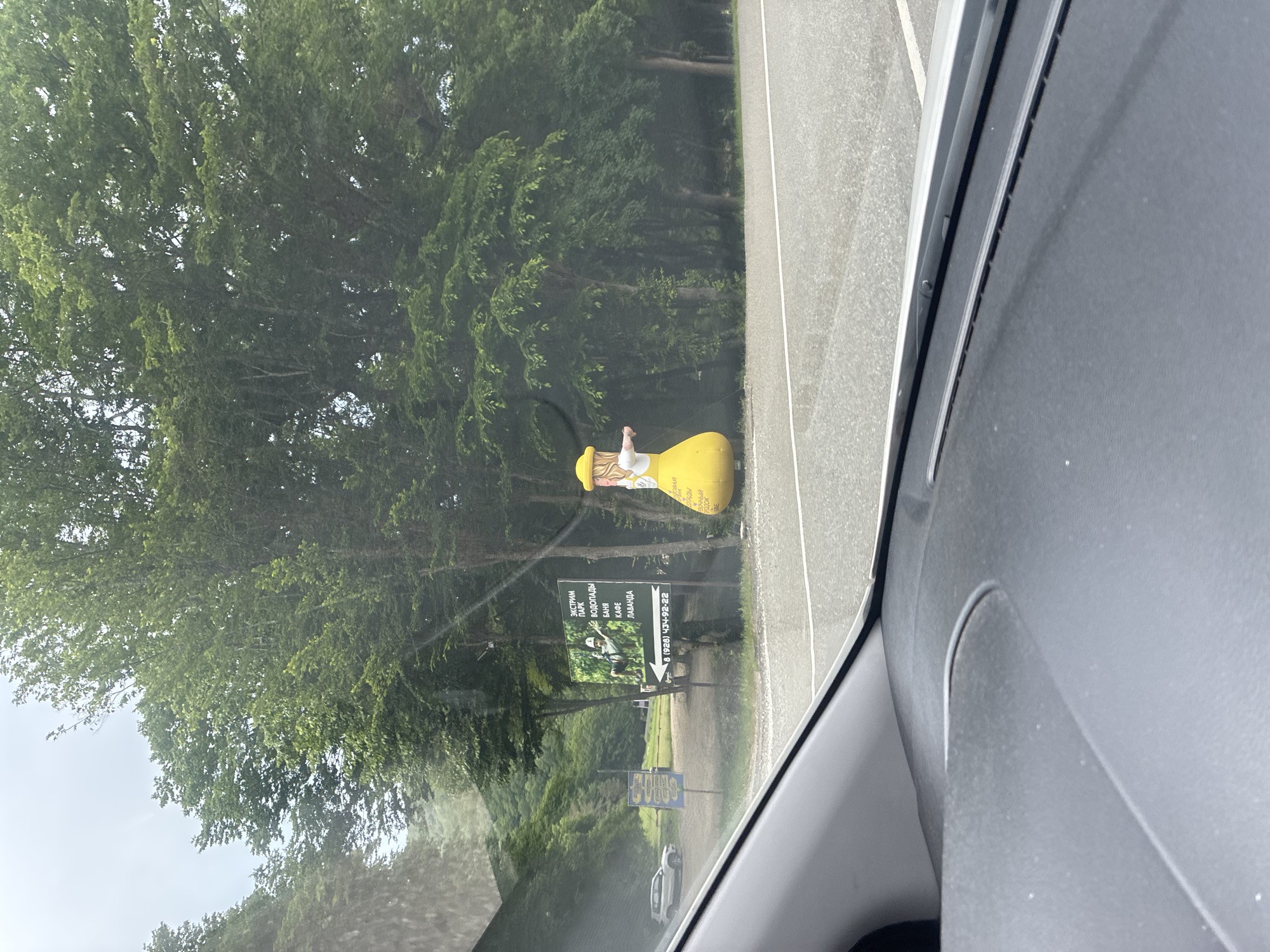In Arkhipo-Osipovka, Russia, a road leads through a wooded area. A large inflatable figure, shaped like a woman in a yellow dress and hat, stands on the side of the road. She has blonde hair and is gesturing with her arms. A signpost with Russian text and an image of a person on a zip line is visible nearby, along with another smaller blue sign. A white SUV is parked on the left side of the road. The scene appears to be captured from inside a vehicle, with the dashboard visible in the foreground. The sky is overcast.