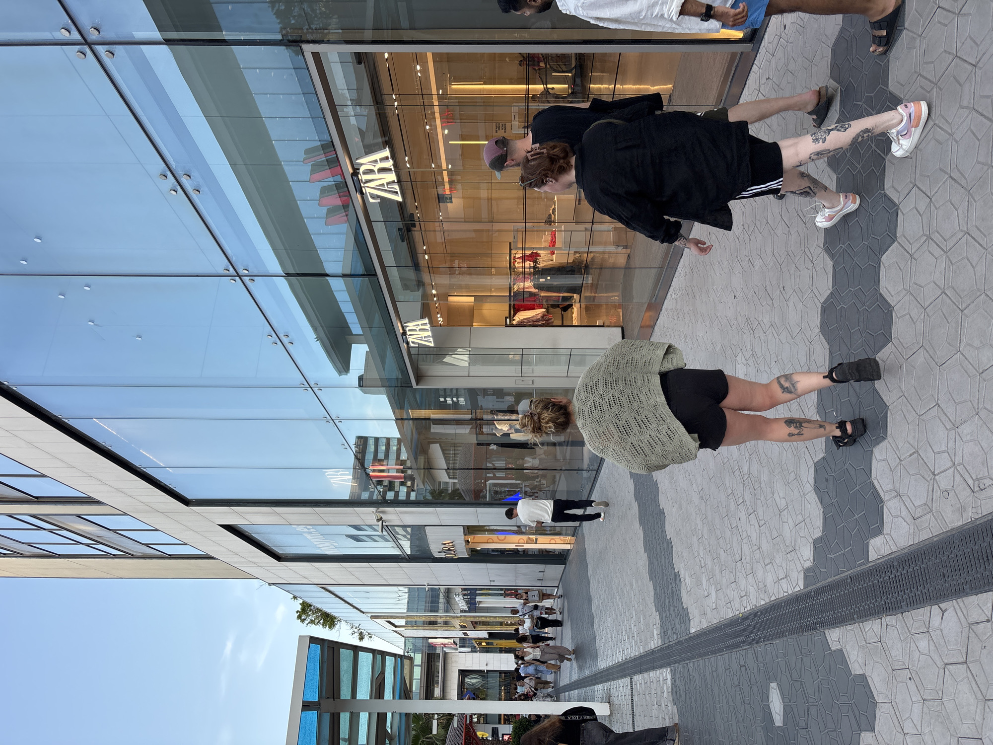 This image captures a bright, daytime scene in a modern pedestrian shopping area, likely a promenade in Barcelona, Spain. The perspective is tilted, showing the ground plane and tall glass-fronted buildings stretching upwards.
In the mid-ground, two individuals are prominent. One person, seen from the back, wears a black long-sleeved shirt, black shorts with white stripes, and colorful sneakers (white with pink and orange accents). They also wear a purple baseball cap and have numerous tattoos visible on their legs. Walking slightly behind them to the left is another person, also seen from the back, wearing a light green or khaki open-knit sweater over black shorts and black sandals. This individual also has visible leg tattoos.
Further to the right, partially cut off at the top, are two men. One wears a white shirt and light blue shorts with black sandals, while the other wears a dark shirt and dark shorts. In the background, numerous other people are scattered along the wide paved walkway, some walking, others appearing to be browsing or relaxing.
The architecture consists of tall buildings with extensive glass storefronts that reflect the clear blue sky and surrounding structures. The prominent store "ZARA" is clearly visible on the glass, both directly and in reflection, indicating a retail environment. Further down the street, "PANDORA" can also be seen on another storefront. The ground is paved with a distinct geometric pattern in varying shades of grey, and a long metal grate, likely for drainage, runs along the center of the walkway. In the lower left, a building features blue-tinted glass panels. The overall impression is a bustling urban setting under clear, sunny skies. A partial reflection of "Y LOLA B" is visible in the bottom right corner.