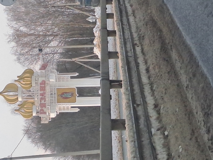 The image displays an outdoor scene depicting the entrance to the city of Ryazan, Russia, observed from a roadside vantage point near Turlatovo. A prominent white welcome sign for Ryazan stands centrally, featuring three golden, onion-shaped domes atop its structure. Below the domes, the city name "РЯЗАНЬ" is displayed in bold red Cyrillic letters. Beneath the text, a shield-shaped emblem depicts a standing figure, likely a warrior or prince, holding a sword.
In the foreground, a grey, multi-segment metal guardrail extends horizontally across the lower portion of the frame, separating the viewer from the area immediately surrounding the sign. The ground beside the road and behind the guardrail is covered with dirty, patchy snow and exposed brown earth. A paved asphalt road surface is visible in the lower right, partially obscured. The background consists of a dense line of bare deciduous trees under a light, possibly overcast, sky. Two utility poles are visible: one to the left of the sign and another to its right. A portion of the dark-colored cabin of a truck is visible on the far right edge of the image. No individuals are present within the scene.