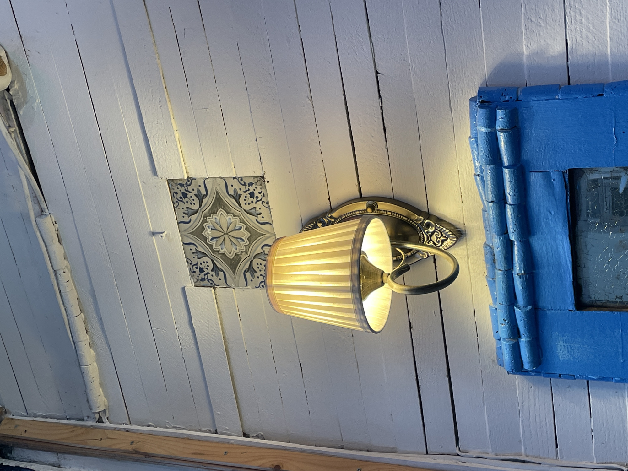 The image depicts an interior scene in Tbilisi, Georgia, likely within a residence. The focal point is a wall lamp with a pleated shade, emanating a warm glow. The lamp is mounted on a wall constructed of white-painted wooden planks. Adjacent to the lamp is a decorative ceramic tile featuring a floral pattern. To the right of the lamp is a blue-painted window frame, partially obscuring a view. The overall impression is that of a cozy, eclectic, and somewhat rustic interior.
