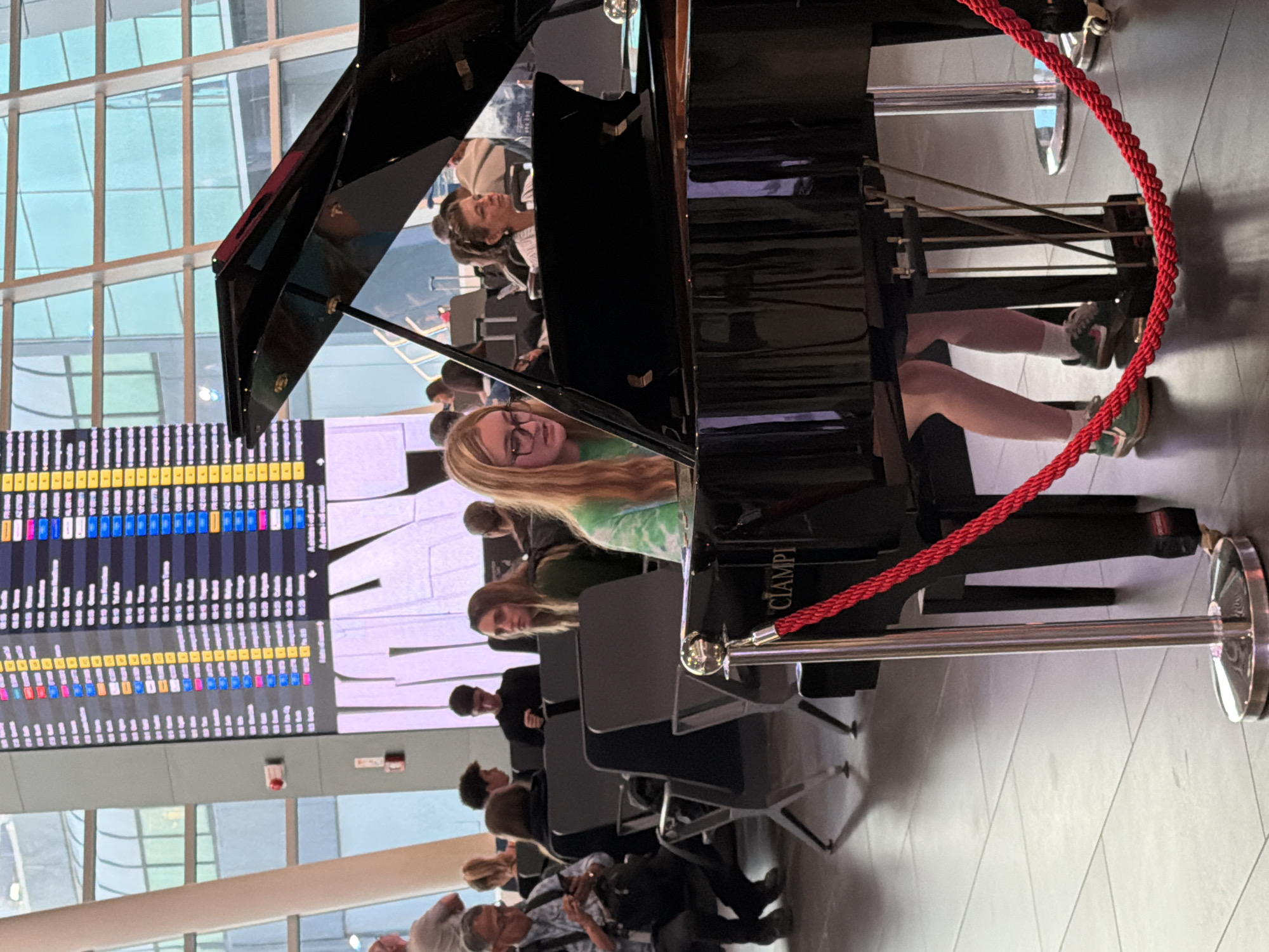 The image depicts a young woman playing a grand piano in an airport terminal. She is seated at the piano, wearing glasses, a green tie-dye shirt, and athletic shoes. Behind her, other travelers are seated in chairs, some looking towards the piano and others engaged with their phones or looking away. In the background, a large digital flight information board displays various destinations and flight details. The environment appears to be a modern airport, with large windows letting in natural light and polished floors. The piano is partially covered by a red rope barrier. The text "CIAMPI" is visible on the side of the piano. This scene is likely at Fiumicino Airport Leonardo da Vinci International Airport in Fiumicino, Italy, given the presence of an airport terminal with flight information boards and a piano performance setting, a common feature in some major international airports.