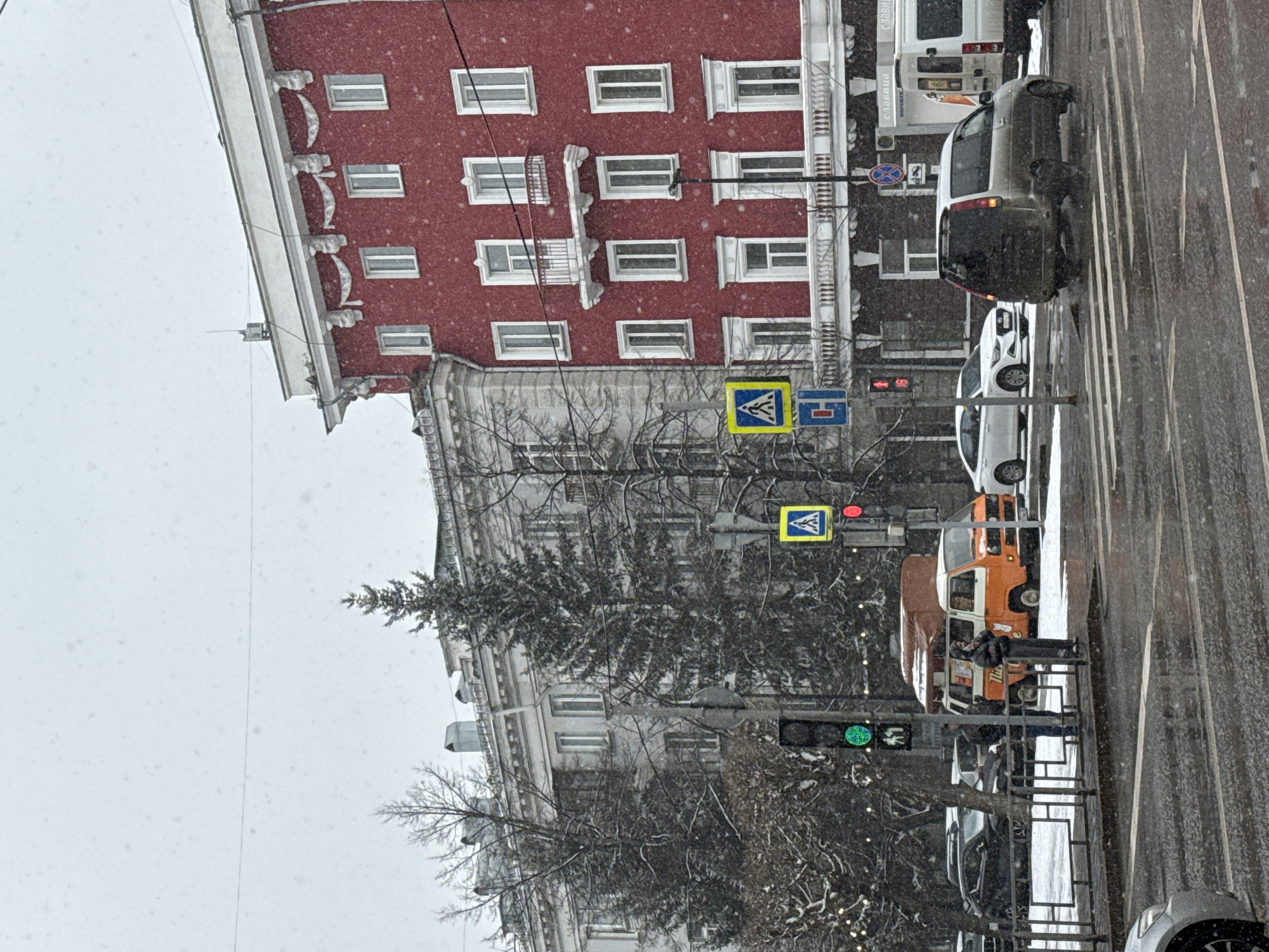 This image captures a snowy street scene in Krasnoyarsk, Russia, oriented 90 degrees clockwise. It depicts a multi-lane urban road flanked by buildings and trees, with visible snowfall under an overcast sky, suggesting daytime.
A prominent red building with numerous white-framed windows and decorative white balconies and cornices stands on the left side of the frame. Below it, a lighter, possibly grey, building with similar architectural details is partially visible through bare tree branches and an evergreen tree.
On the street, a zebra crossing is visible. Several vehicles are present: a dark SUV, a white sedan, a distinctive orange/brown vintage-style van, and a white commercial van. At least two pedestrians are seen on the sidewalk near the orange van, with one leaning against a black metal railing.
Traffic infrastructure includes two yellow diamond-shaped pedestrian crossing signs, a blue rectangular sign (possibly indicating a parking or stopping zone), and traffic lights. One traffic light shows a red signal, while another lower light displays green. The trees lining the street are mostly bare, lightly dusted with snow.
Visible text includes "СЛАВИЦА" and "Мороженое" (Ice Cream) on the side of the white commercial van, and part of "Тигр" (Tiger) on the orange van. The scene conveys a typical winter day in an urban setting.