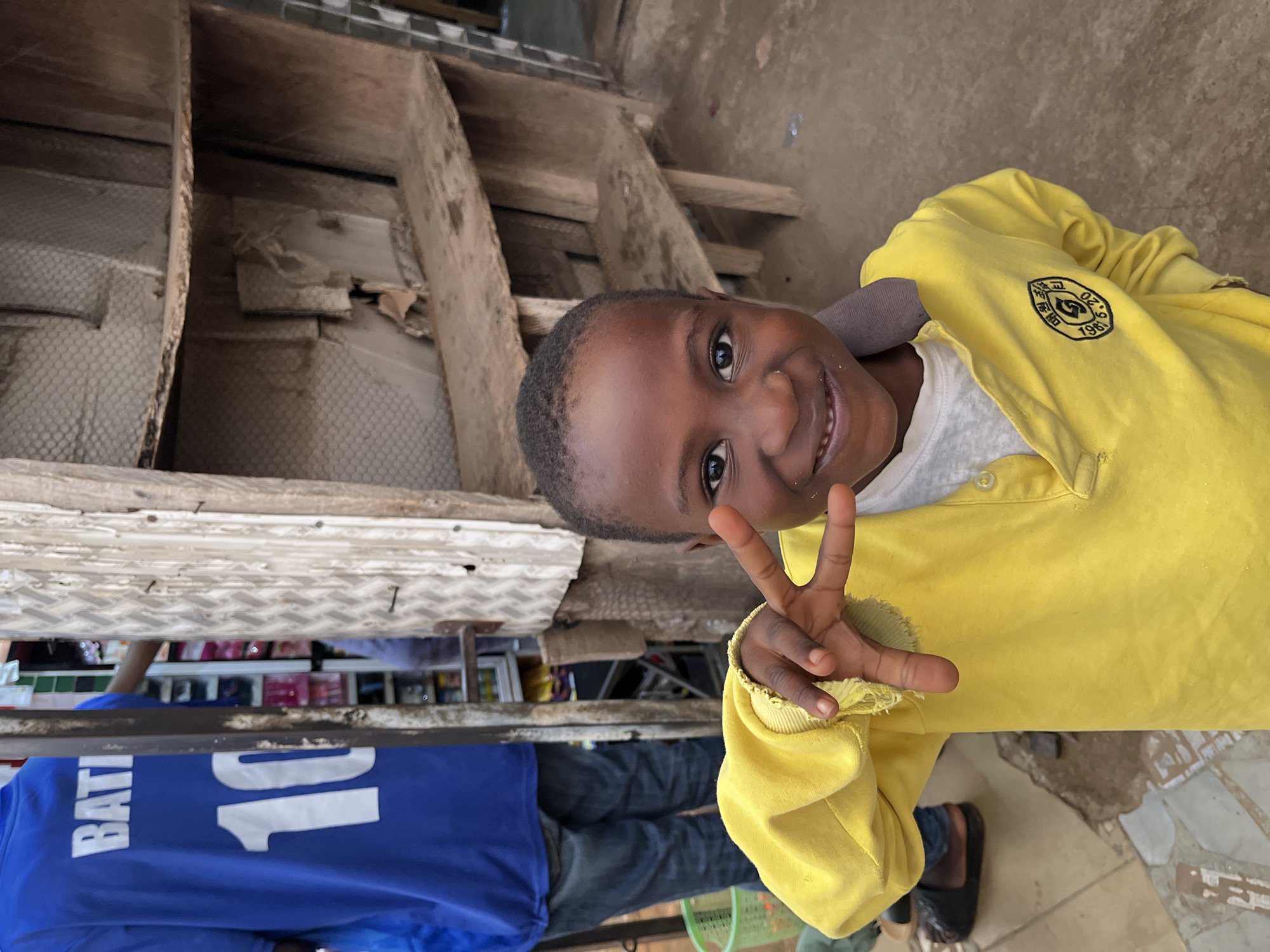 An upright, eye-level shot features a smiling young Black child standing in what appears to be an indoor or semi-sheltered market or shop setting in Jos, Nigeria. The child, facing the camera, makes a peace sign with their right hand, radiating a cheerful and engaging expression. They are dressed in a bright yellow long-sleeved polo shirt over a white undershirt, paired with blue jeans and black slip-on shoes.
On the left chest of the yellow shirt, a circular emblem is visible, containing Korean characters, including "학교" (hakgyo, meaning "school"), and the year "1965".
In the background, a person, likely an adult male, is partially visible from the waist down, wearing a blue t-shirt with "BATI" and the number "10" printed in white on the back, along with blue jeans. Behind them, there are wooden structures, possibly shelving or a counter, and indistinct items on display, suggesting a retail environment. The floor is a mix of concrete and patterned tiles.
The lighting is soft and ambient, indicative of an indoor or shaded location. The overall atmosphere is one of warmth and candid interaction, highlighted by the child's joyful demeanor and direct gaze.