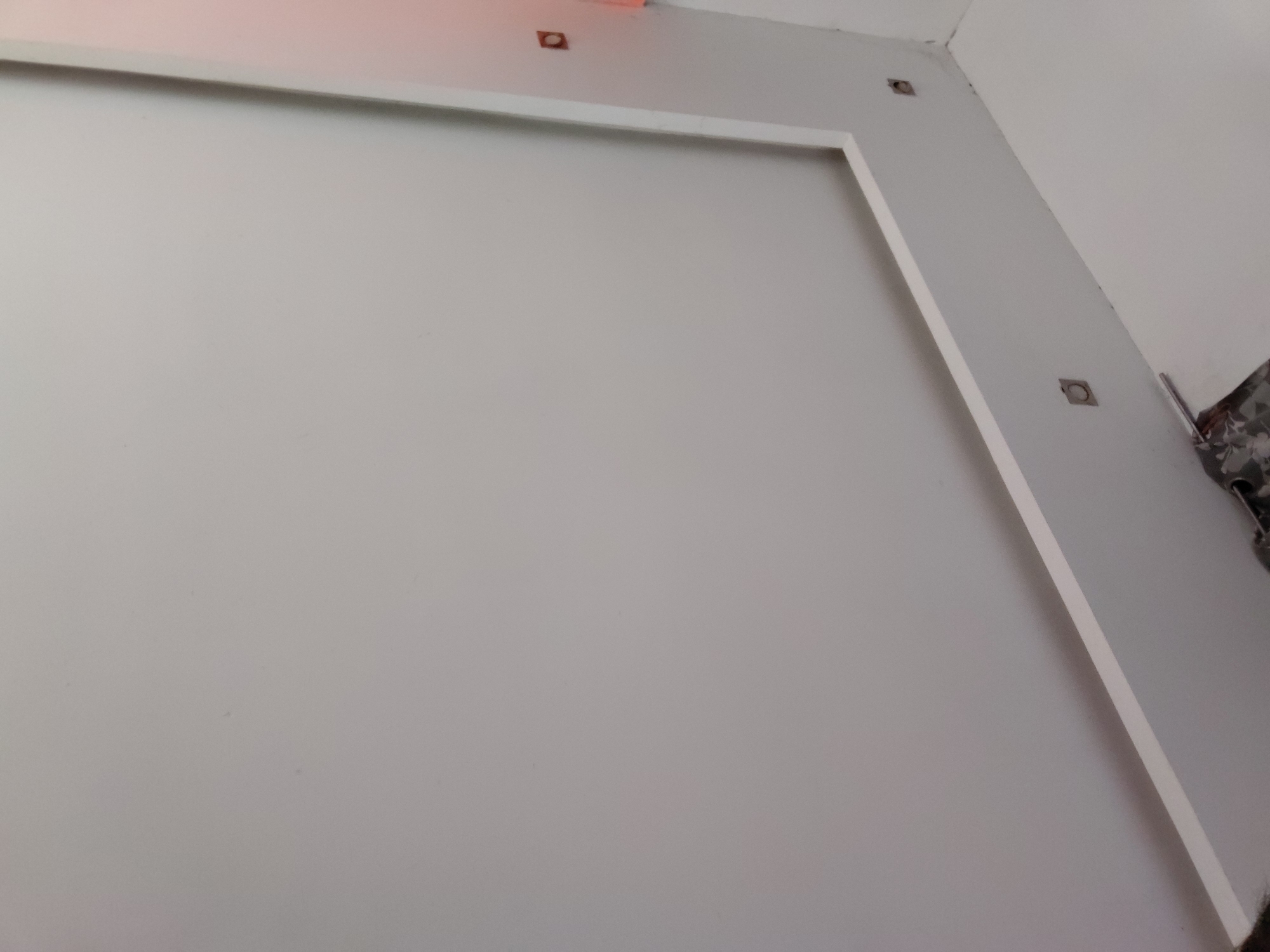 The image captures an interior view of a room, primarily showcasing a large, light-colored wall, likely painted off-white or light grey. A prominent white, rectangular frame or border defines a section of this wall, running along its top and left edges.
Several square-shaped cutouts, appearing dark inside, are visible on the walls. One cutout is positioned near the top left corner of the framed section, with a faint orange-pink glow emanating from just above it, possibly from a light source or reflection. Another similar cutout is situated further to the right along the top edge of the wall. On the right side of the image, two additional square cutouts are present on an adjacent wall, which meets the main wall at a corner, showing some minor surface irregularities. A thin, white vertical strip or molding runs parallel to this corner on the right wall.
In the far right portion of the frame, a crumpled piece of fabric with a patterned design, featuring dark and light tones, is partially visible. This could be a curtain, drapery, or other textile material. The overall scene suggests an indoor setting, possibly a room under construction, renovation, or one that has recently been painted, indicated by the clean but somewhat unfinished appearance and exposed cutouts. There are no people or specific activities depicted. The location context for this scene is Santa Ana, Venezuela.